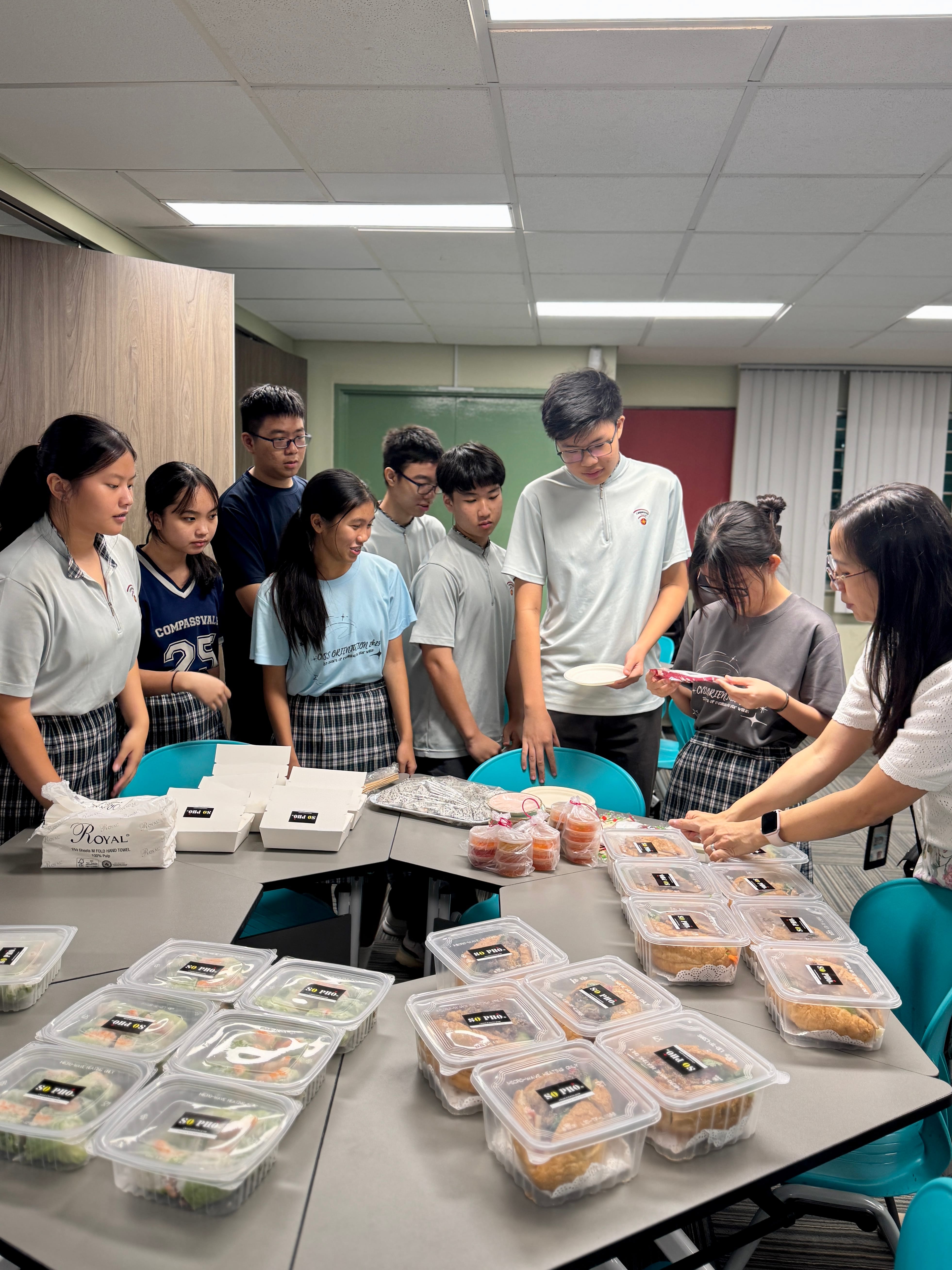 Group of students around tables with stacked takeout meal containers while two women sort items in a classroom.