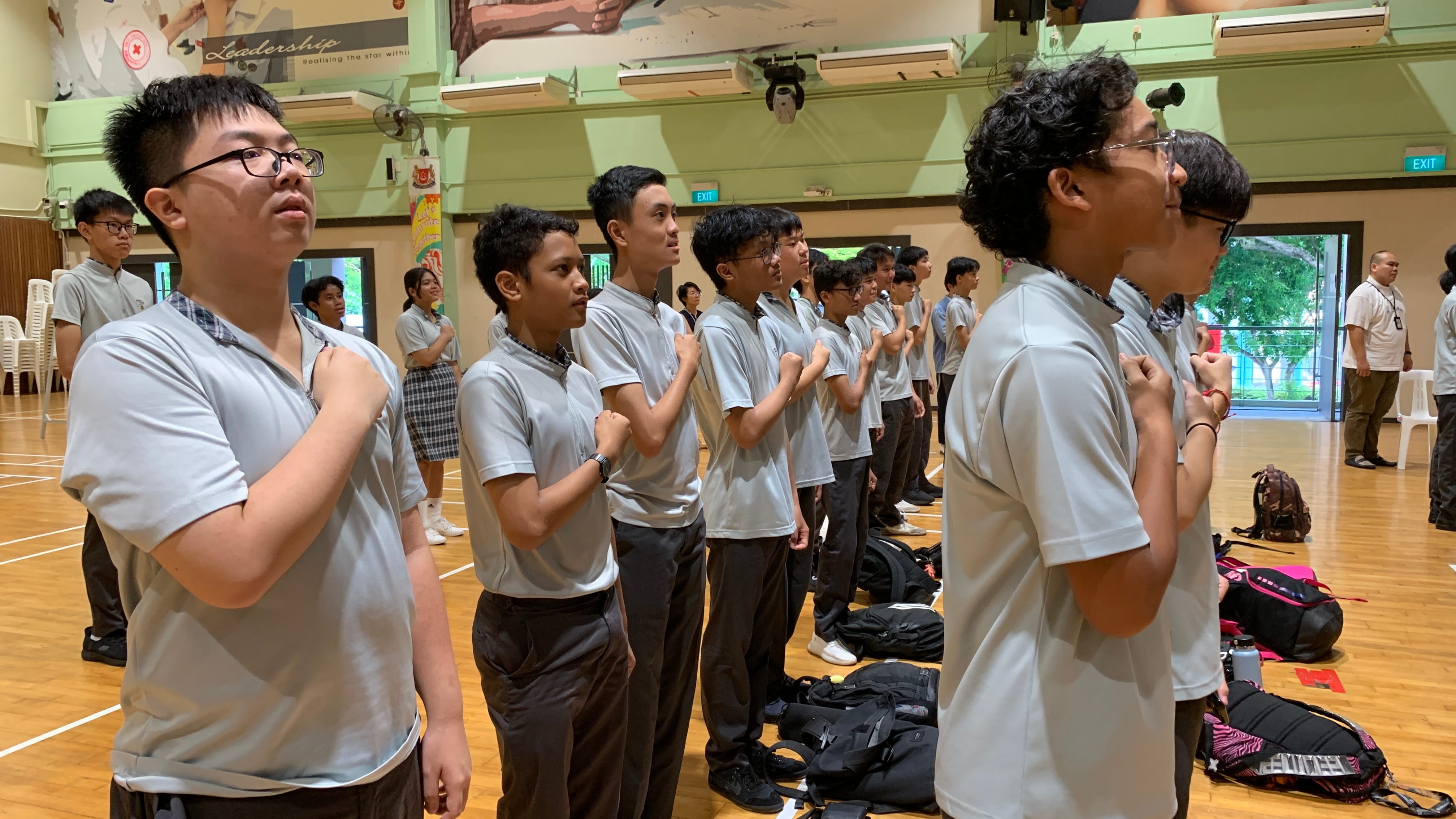 Students in gray uniforms stand in rows in a school hall, fists on chests, reciting a pledge.