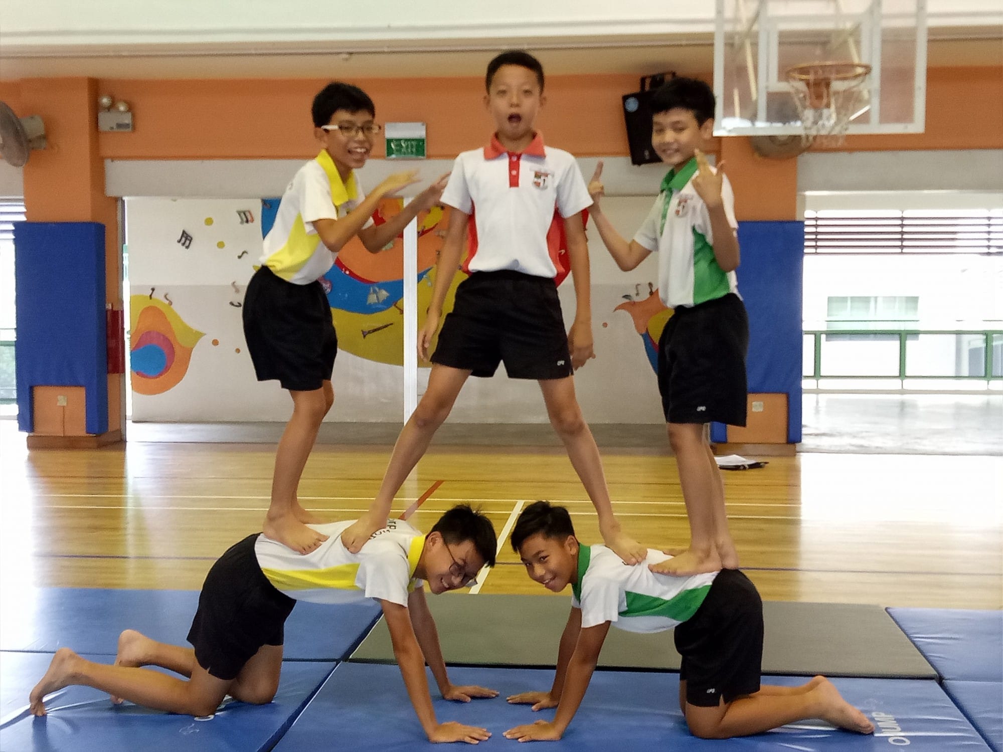 Five boys form a human pyramid on blue gym mats in a school hall, smiling and posing.
