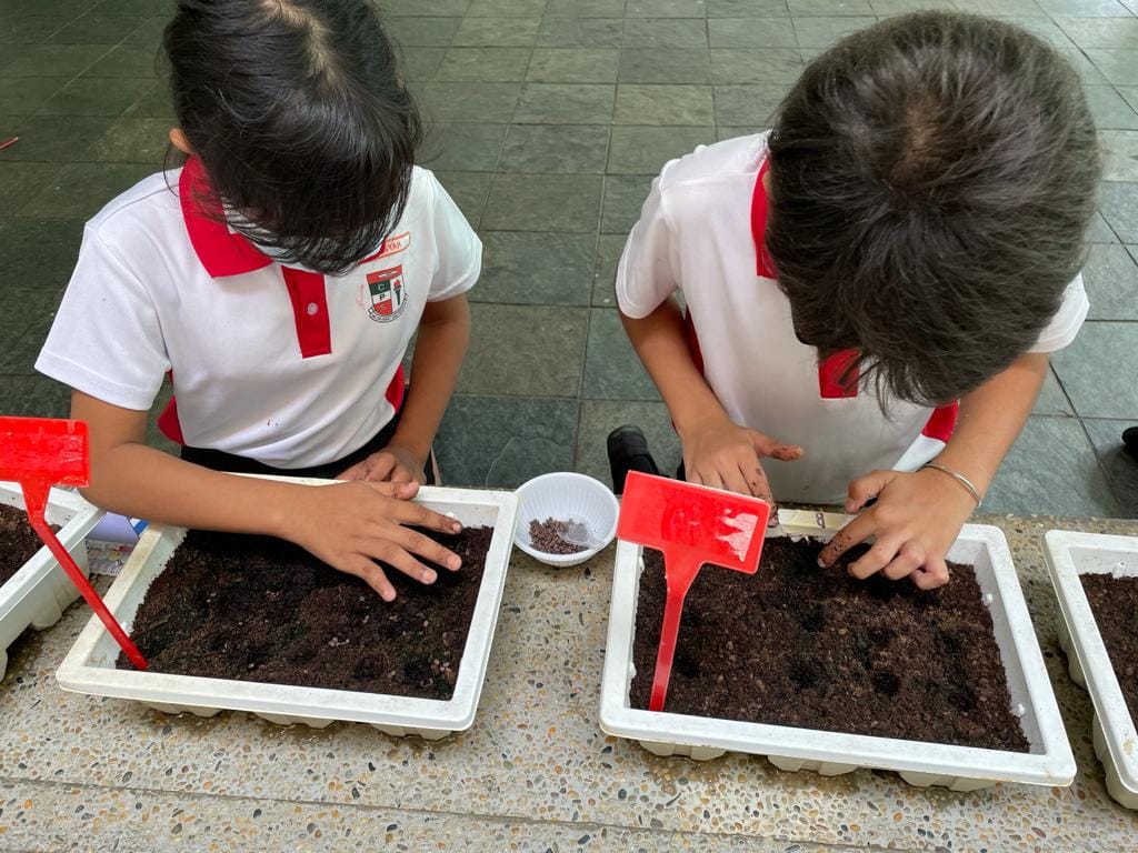 Two school children in white and red uniforms planting seeds in soil-filled trays on a tiled floor.