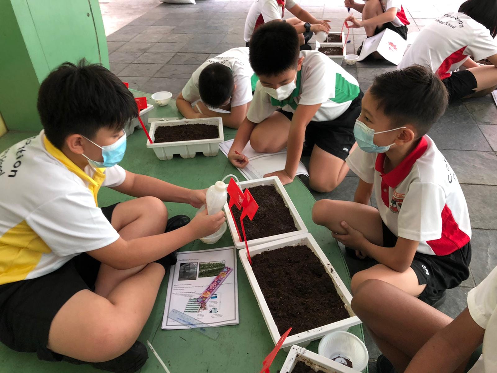 Children wearing masks sit on the floor working with soil-filled trays and worksheets during an outdoor planting activity.