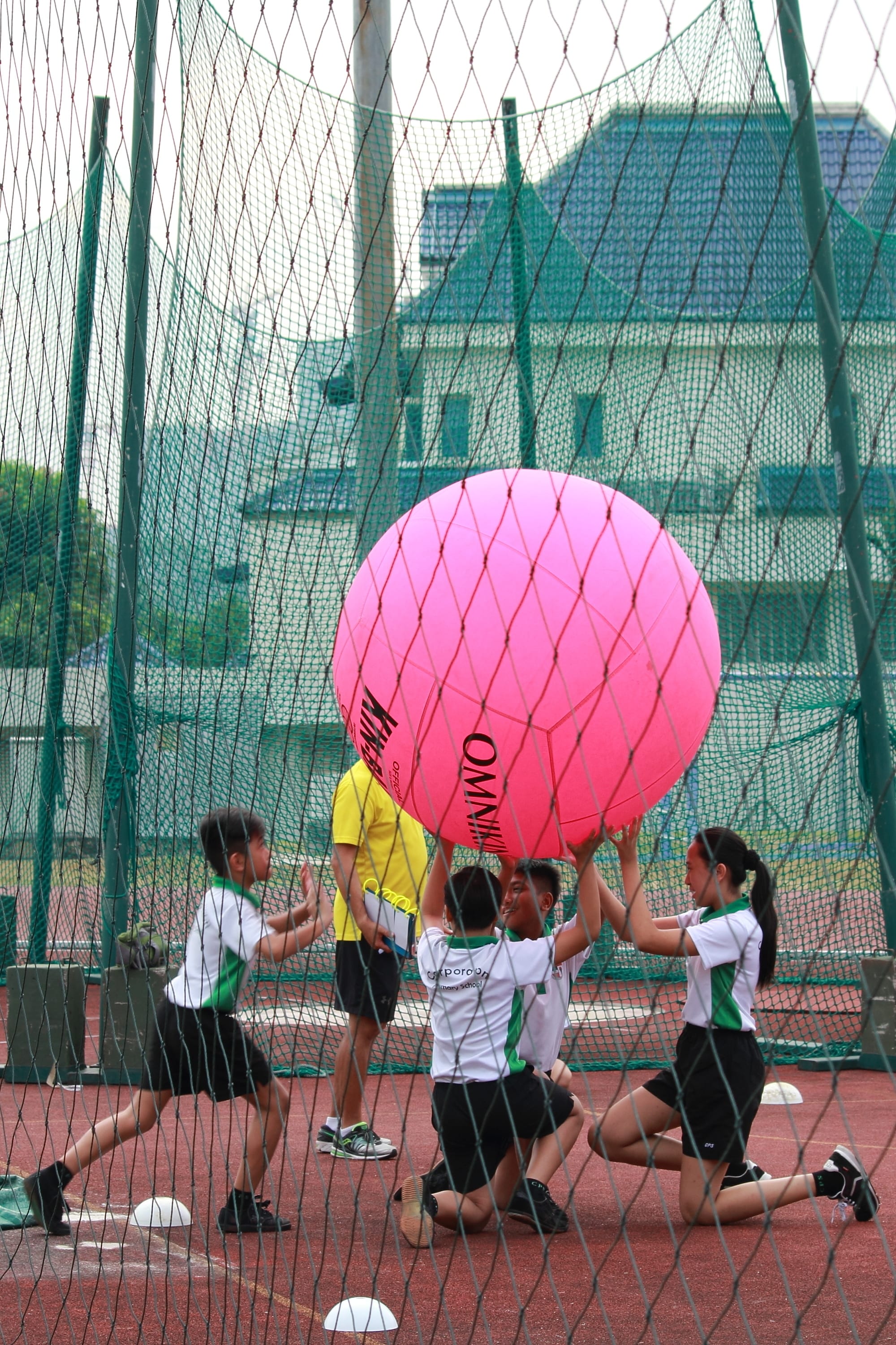 Children in school uniforms lift a large pink ball on an outdoor sports court surrounded by green netting.