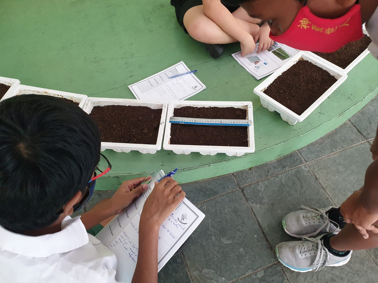 Children sit around a green table with white trays of soil, using rulers and writing on worksheets outdoors.
