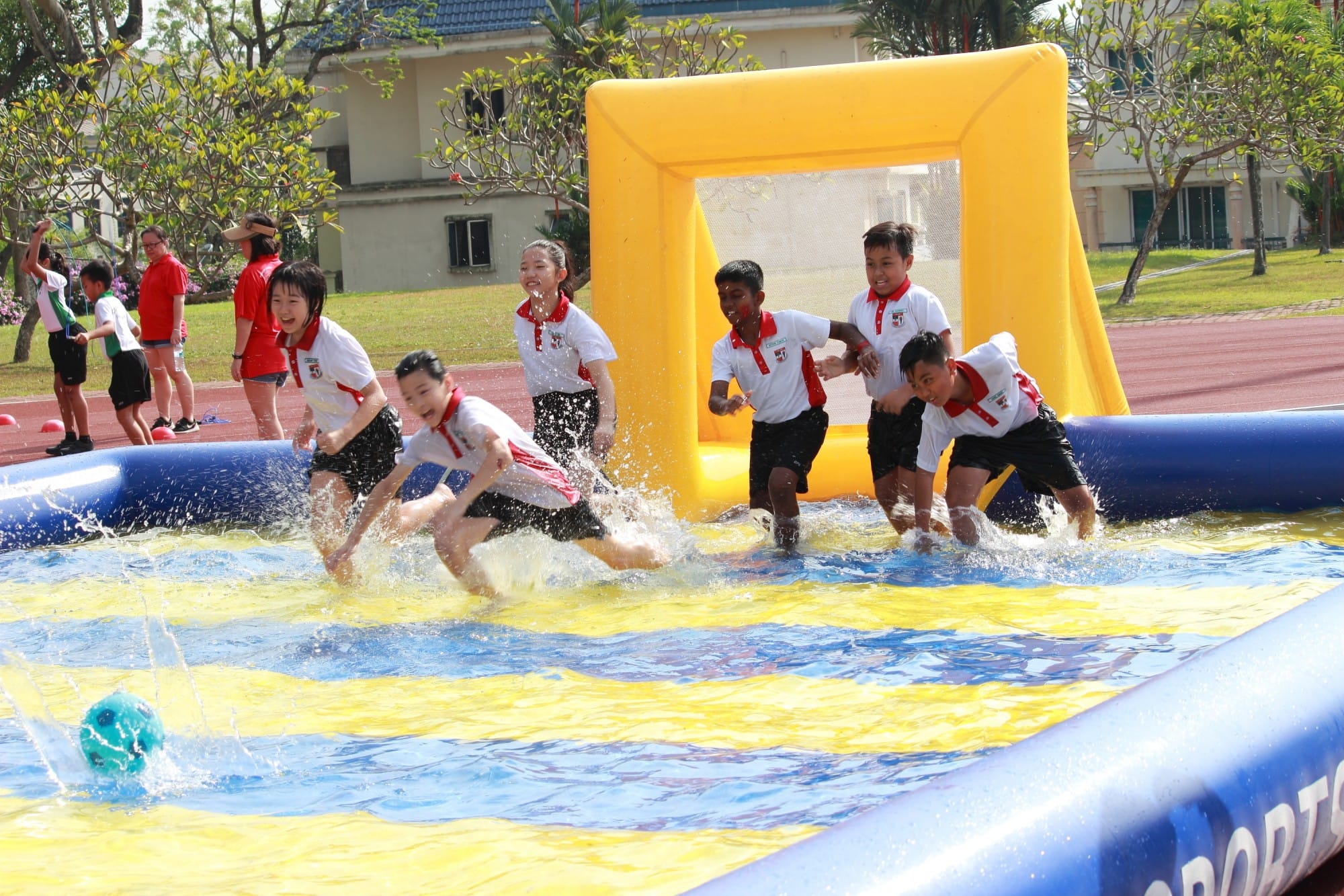 Children in school uniforms play water soccer in an inflatable pool with a yellow goal on a sunny day.