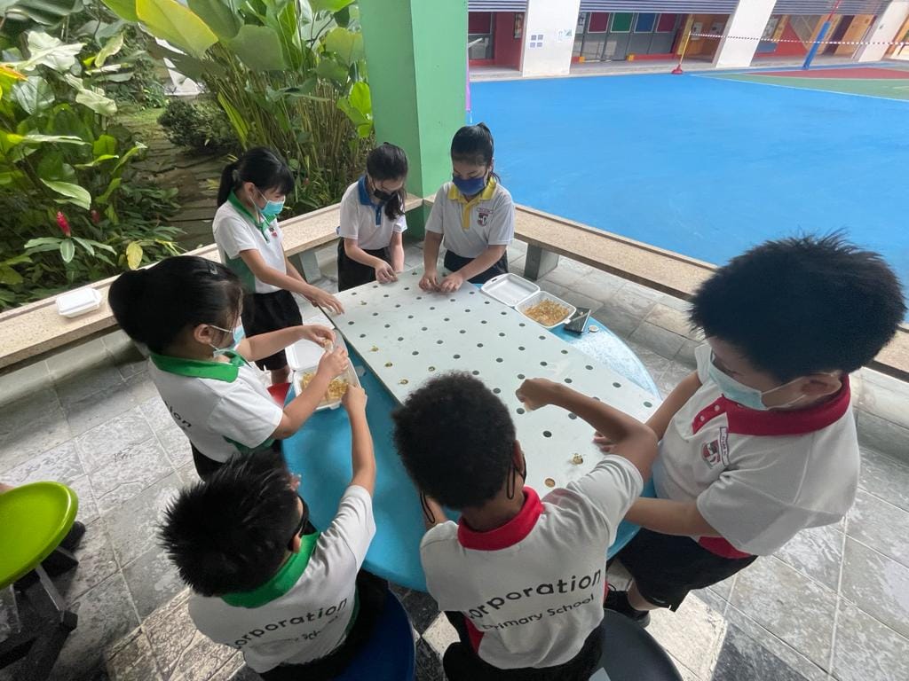 Six masked primary school students in uniforms gather around an outdoor table, placing small objects into holes on its surface.