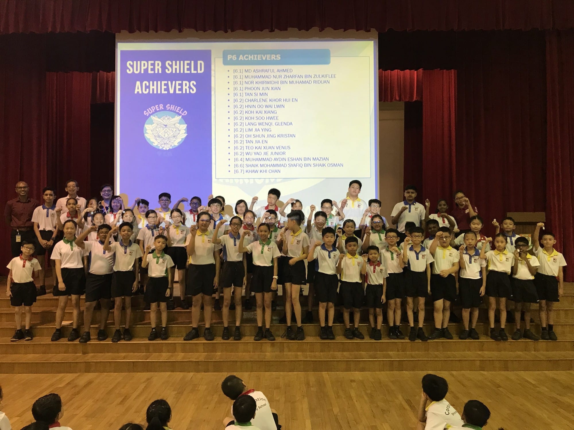A large group of uniformed school students and teachers stand on a stage in front of a Super Shield Achievers presentation screen.