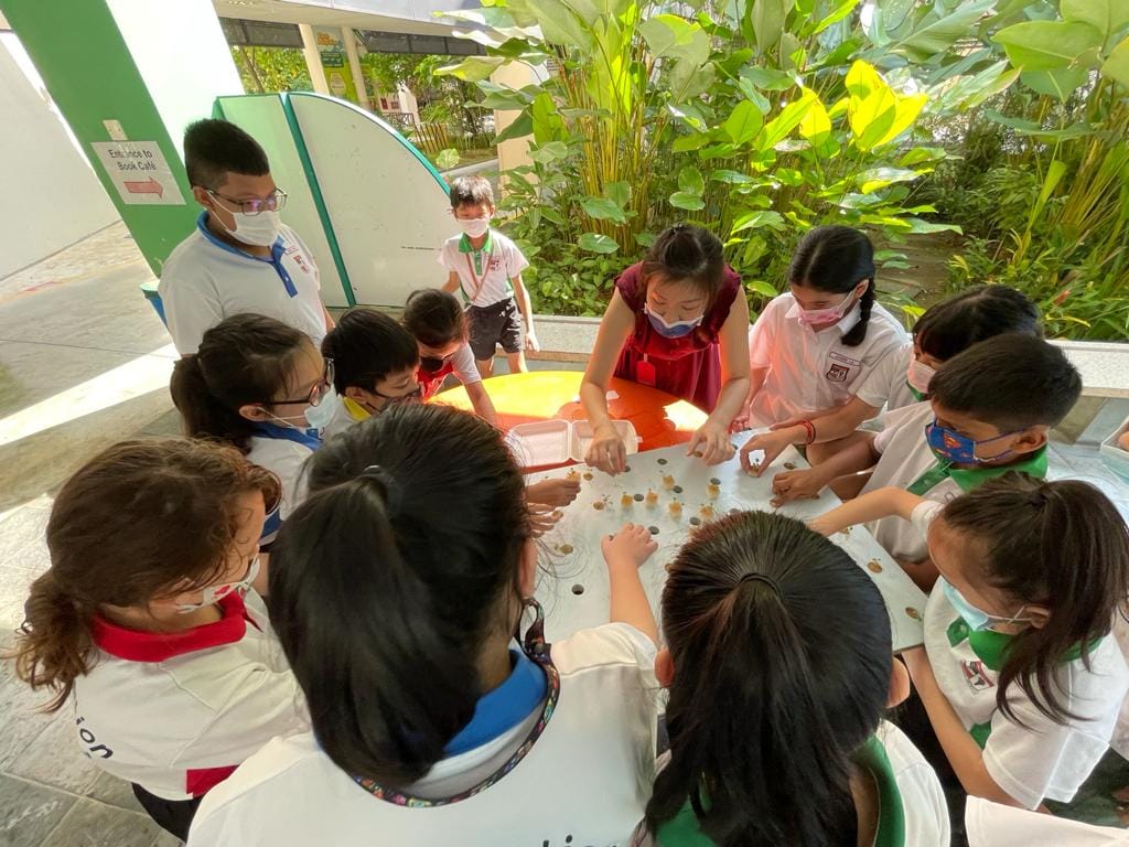 Children wearing face masks gather around an outdoor table, examining small objects scattered on its surface.