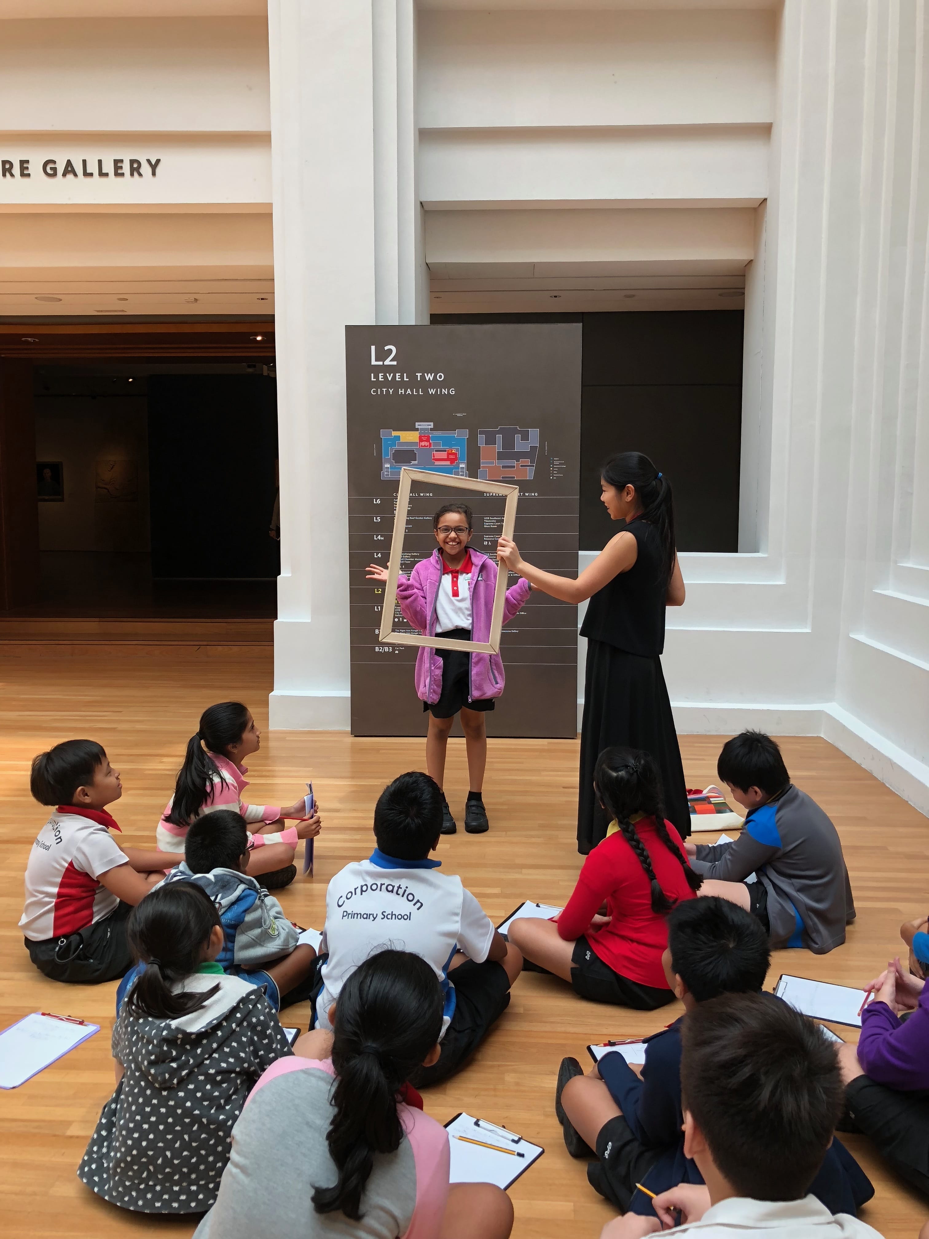 A girl stands inside an empty picture frame held by a teacher while primary school students sit on a gallery floor watching.