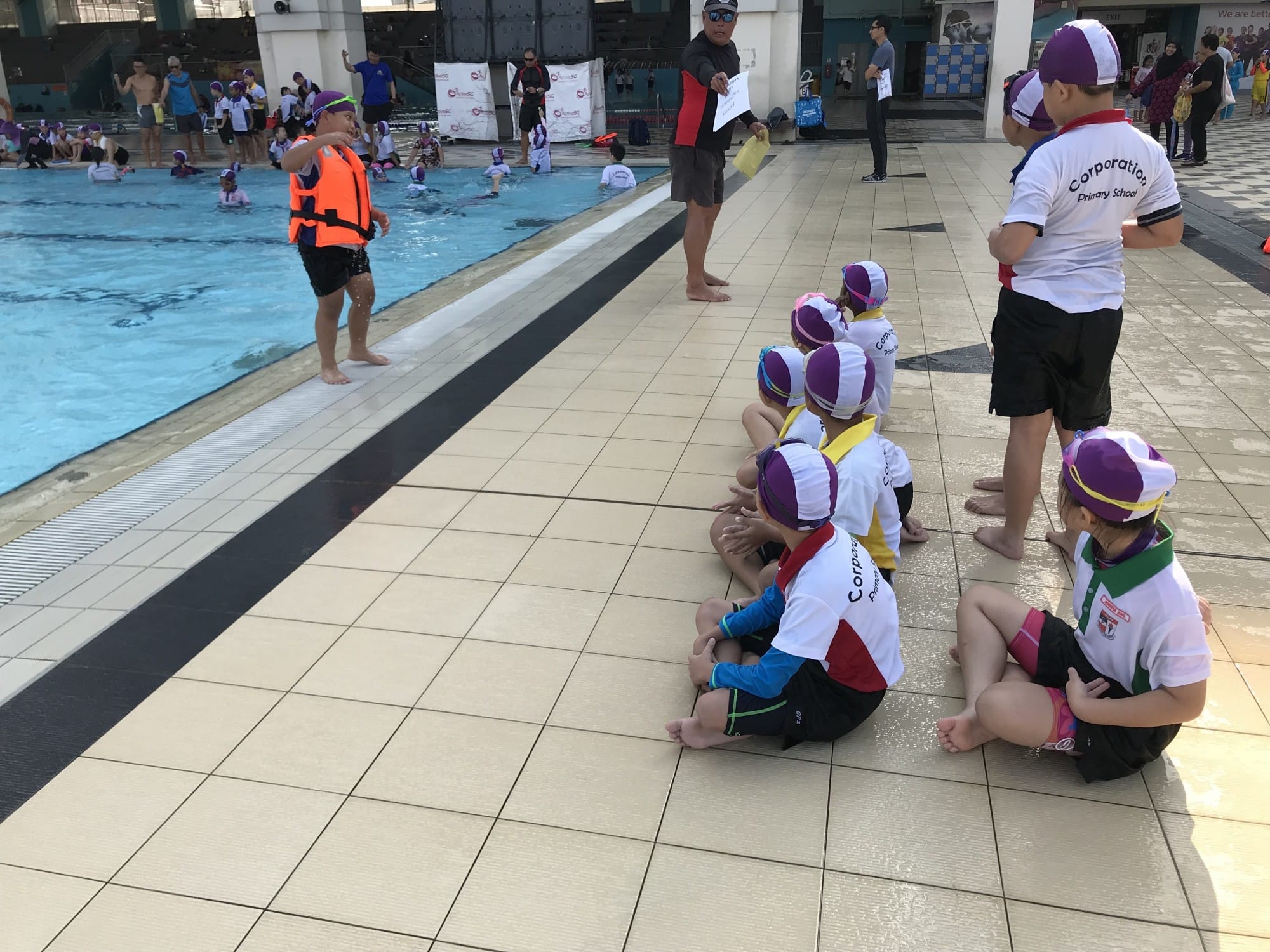Young children in purple swim caps and uniforms sit poolside watching an instructor in an orange life jacket.