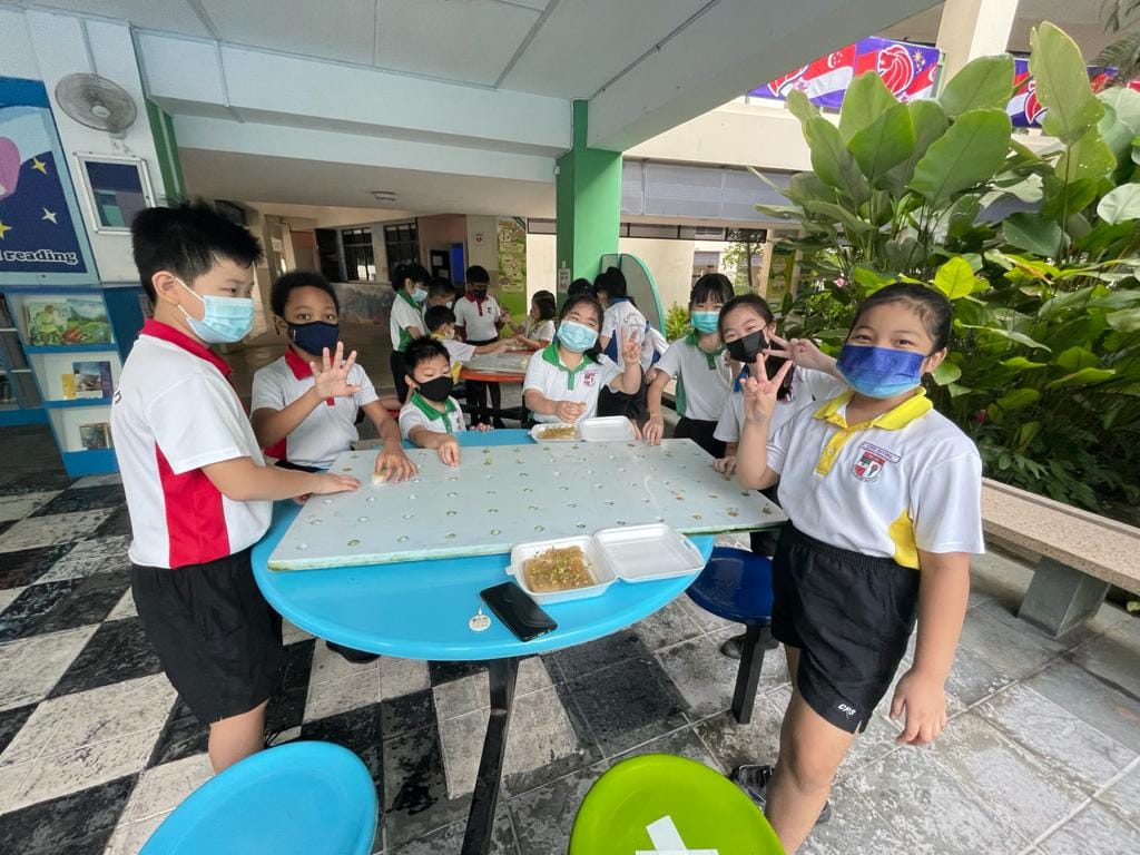 Several young students wearing face masks sit and stand around a blue table with food containers at school.