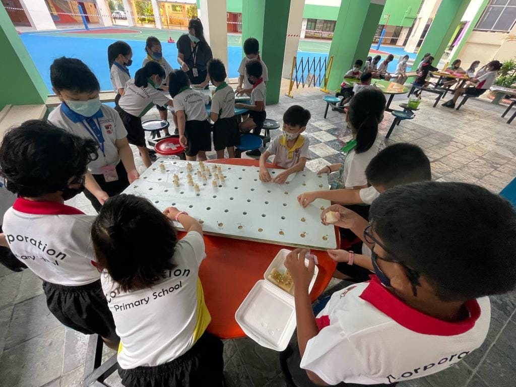Primary school children in uniforms and masks plant seedlings into a white board with holes at outdoor tables.