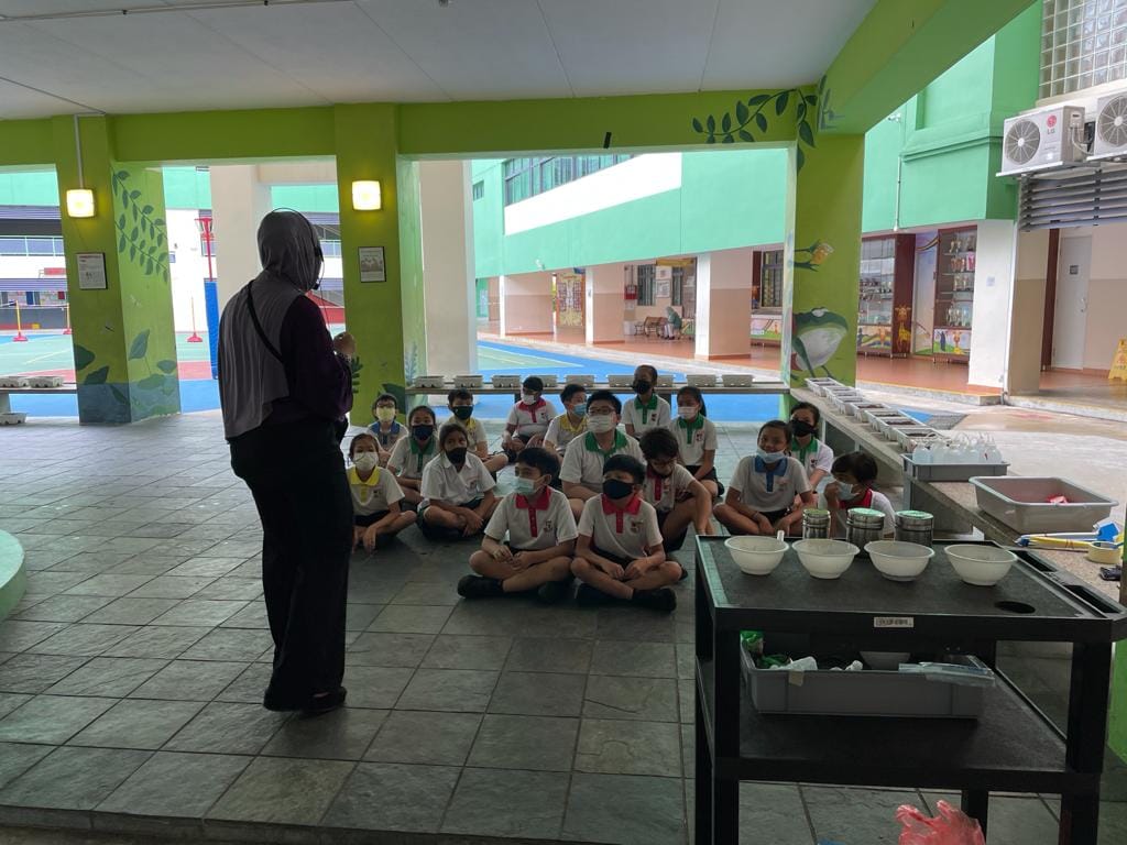 A teacher stands before masked students in uniforms sitting on a floor in a colorful school canteen area.