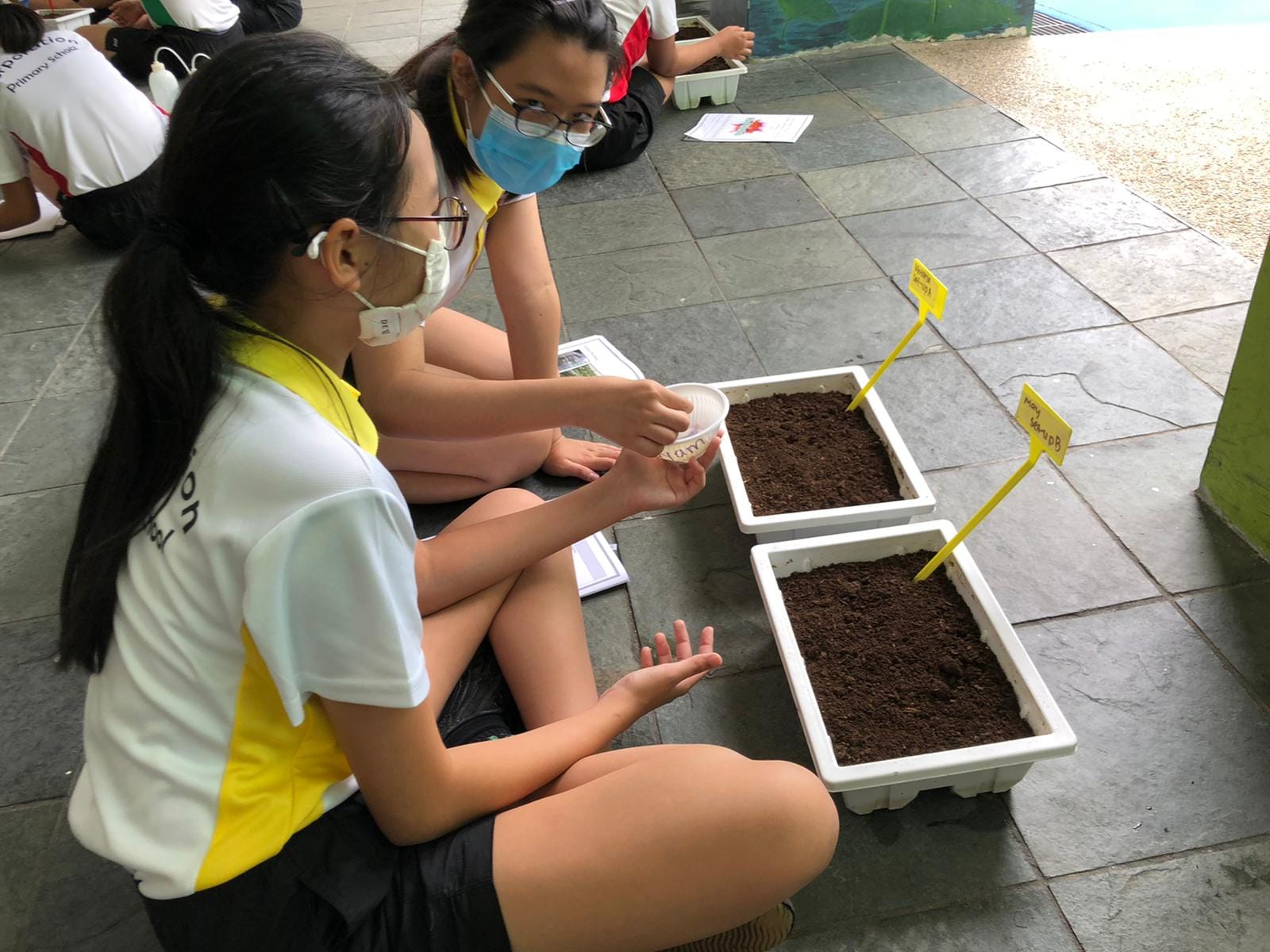 Two masked students sit on a tiled floor planting seeds into soil-filled white trays with yellow label markers.
