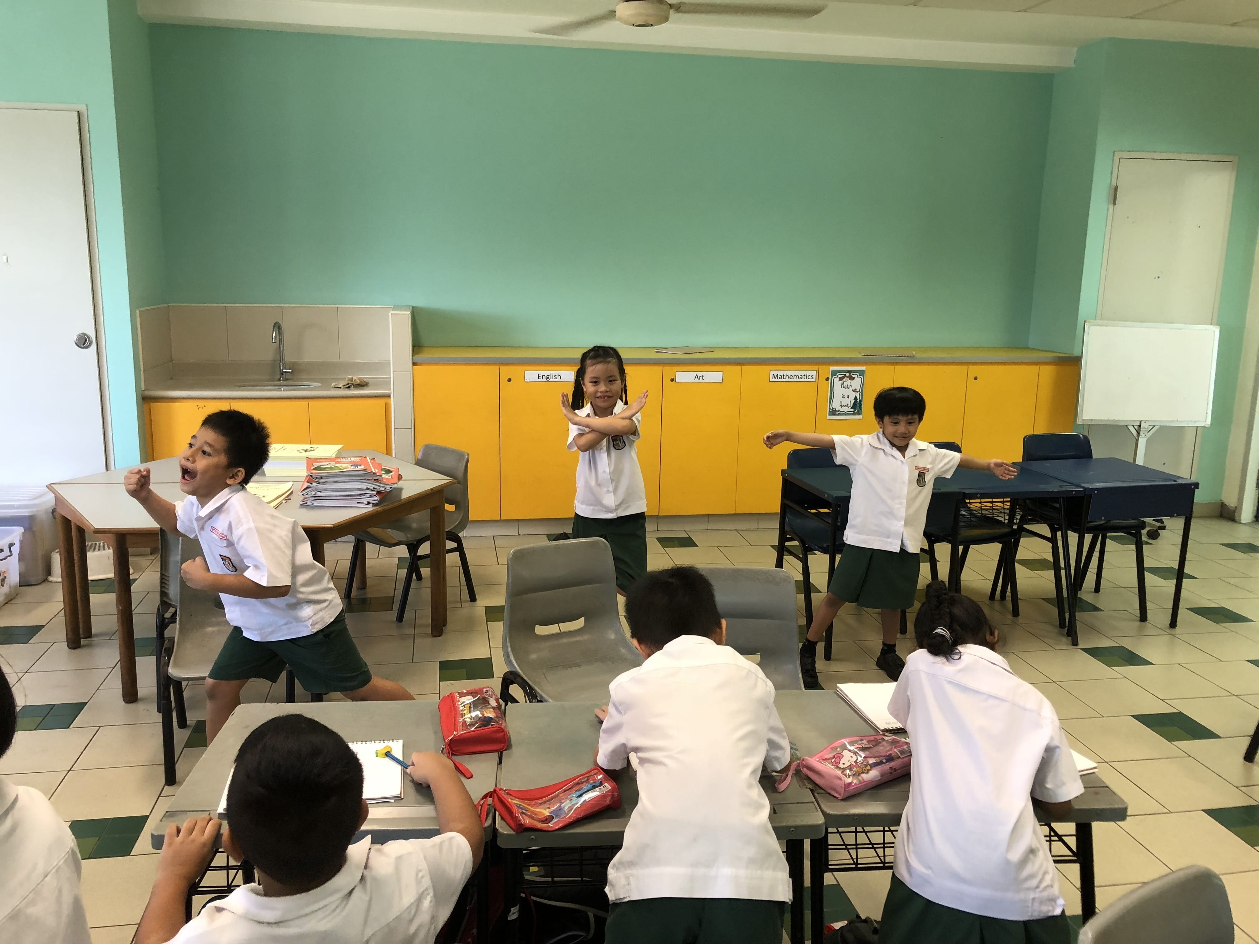 Young students in white uniforms perform an activity in a teal classroom with yellow cabinets and tiled floors.