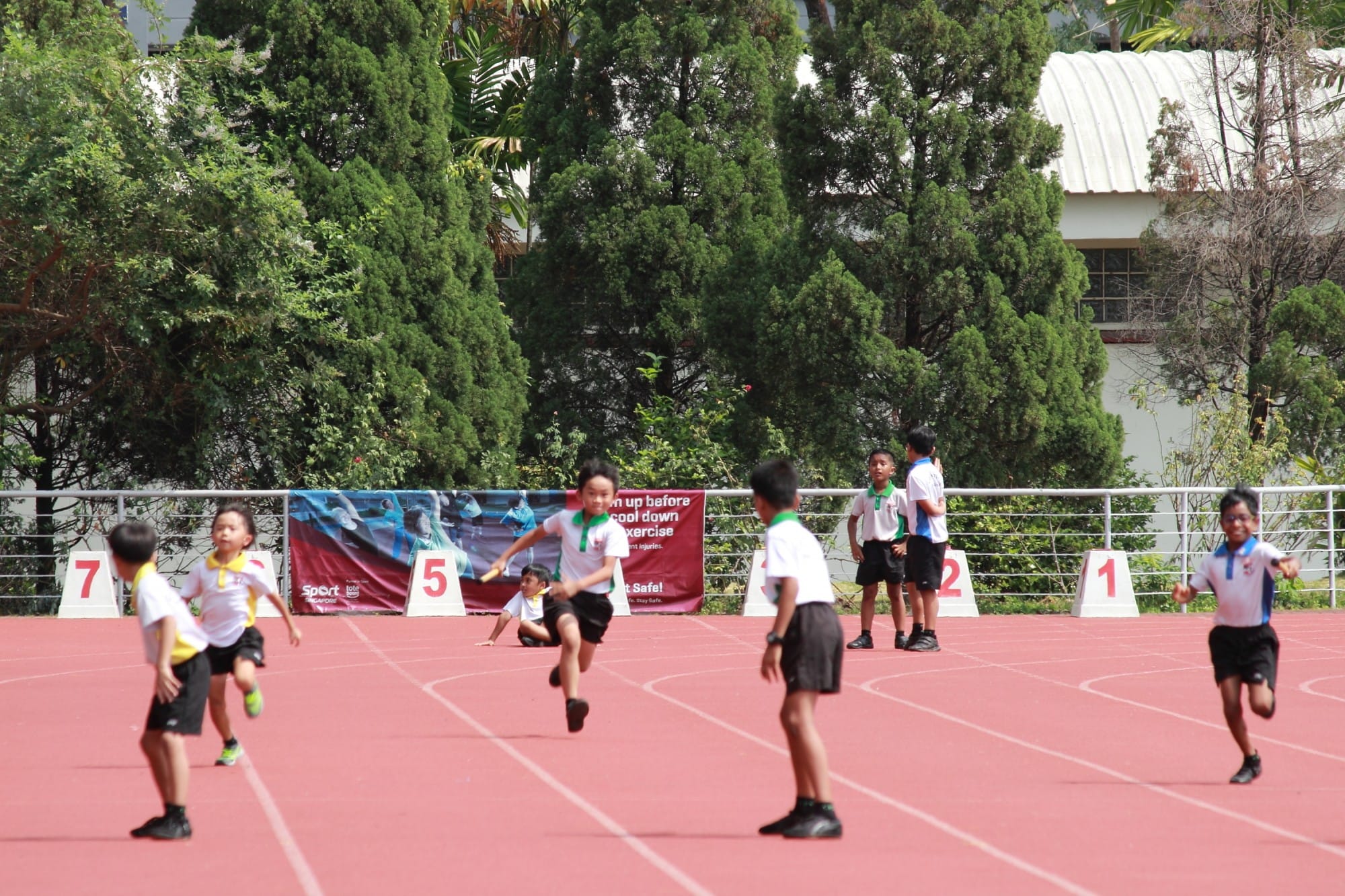 Children in uniforms running a relay race on a red outdoor track with numbered lane markers and trees behind.