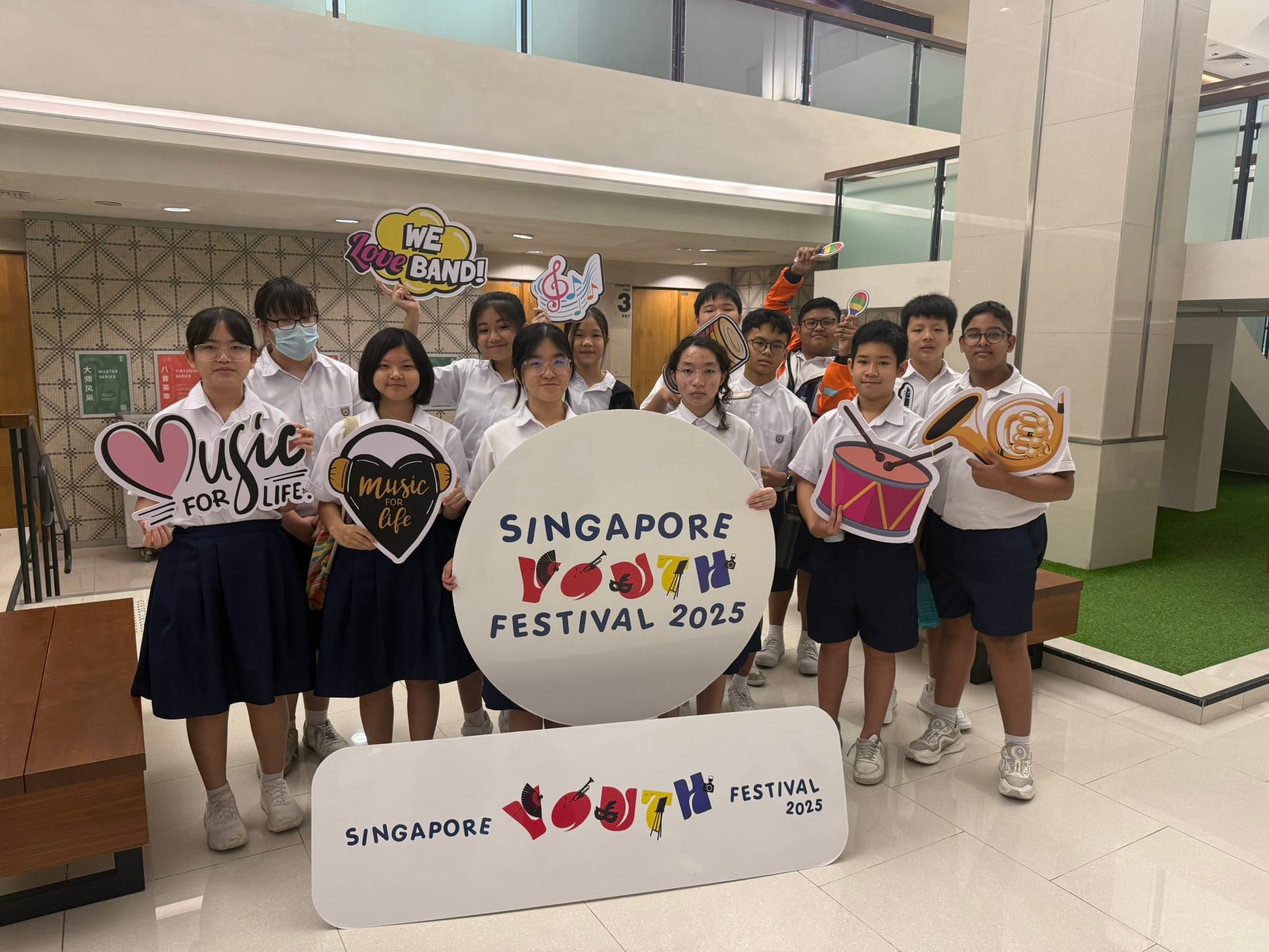 Students in school uniforms pose with Singapore Youth Festival 2025 signs and music-themed props indoors.