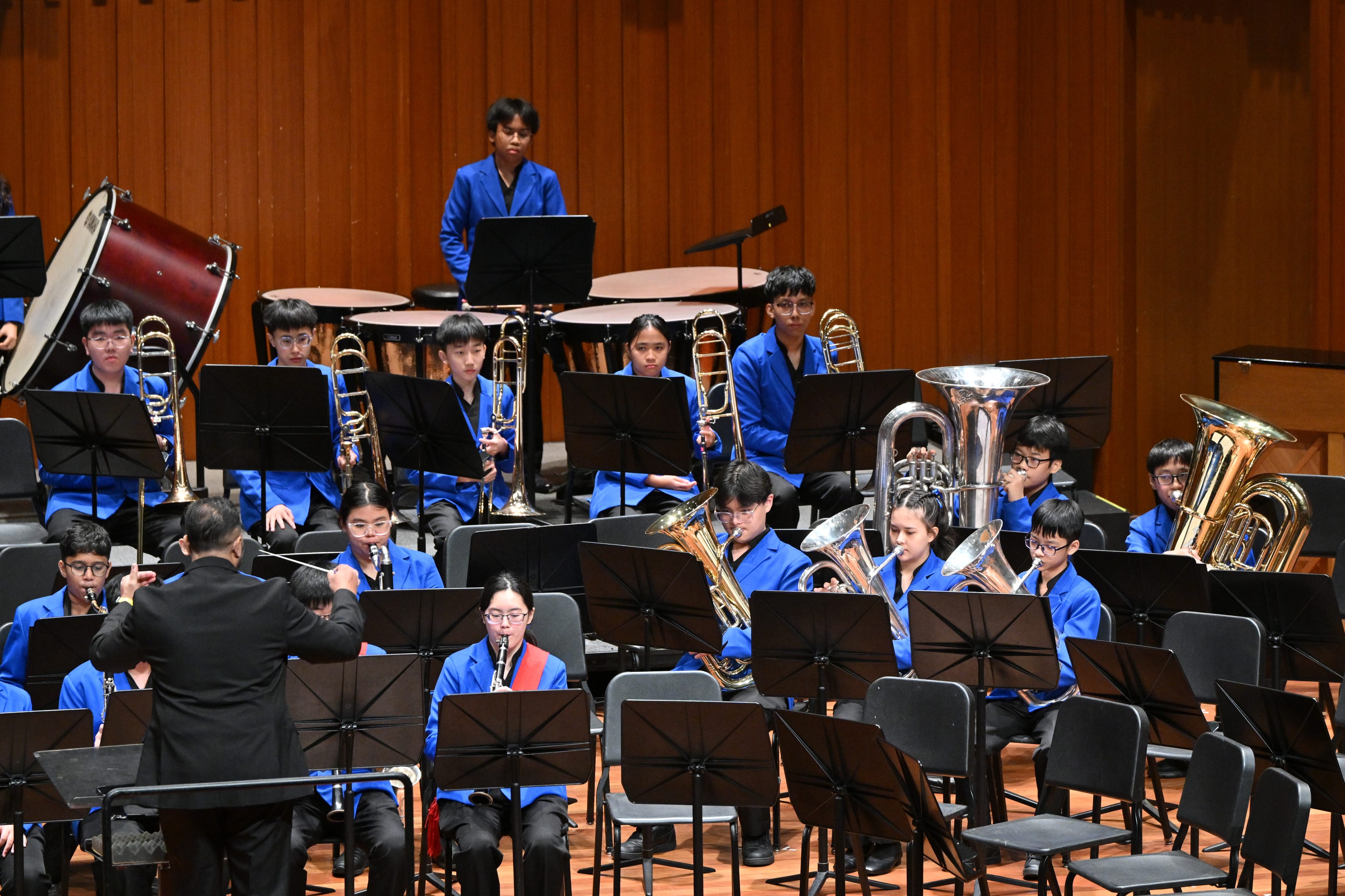 Conductor leads a student band in blue jackets, with brass, woodwinds, and drums on a stage.