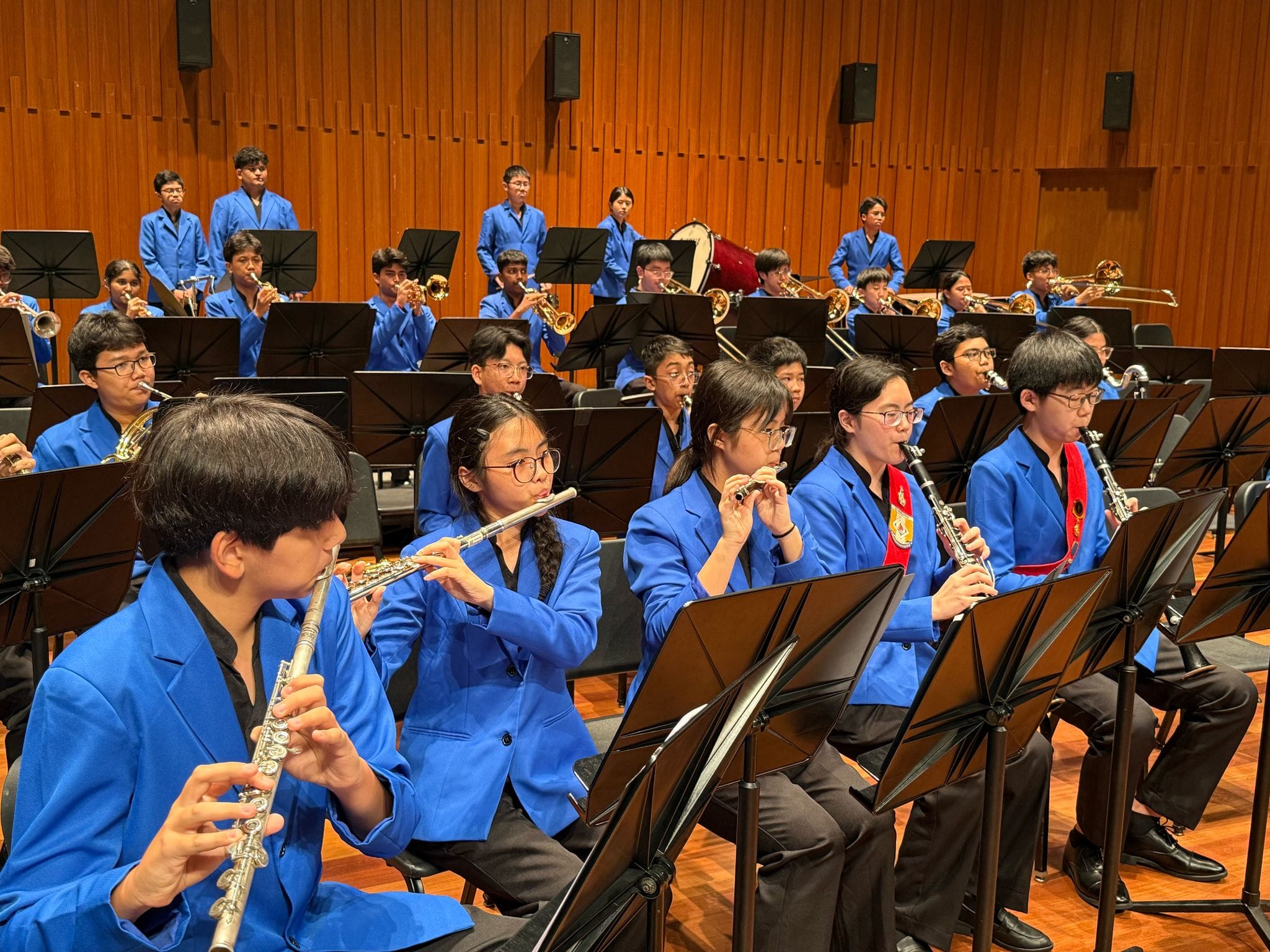 A youth wind band in blue blazers plays flutes, clarinets, and brass instruments on a wooden concert stage.