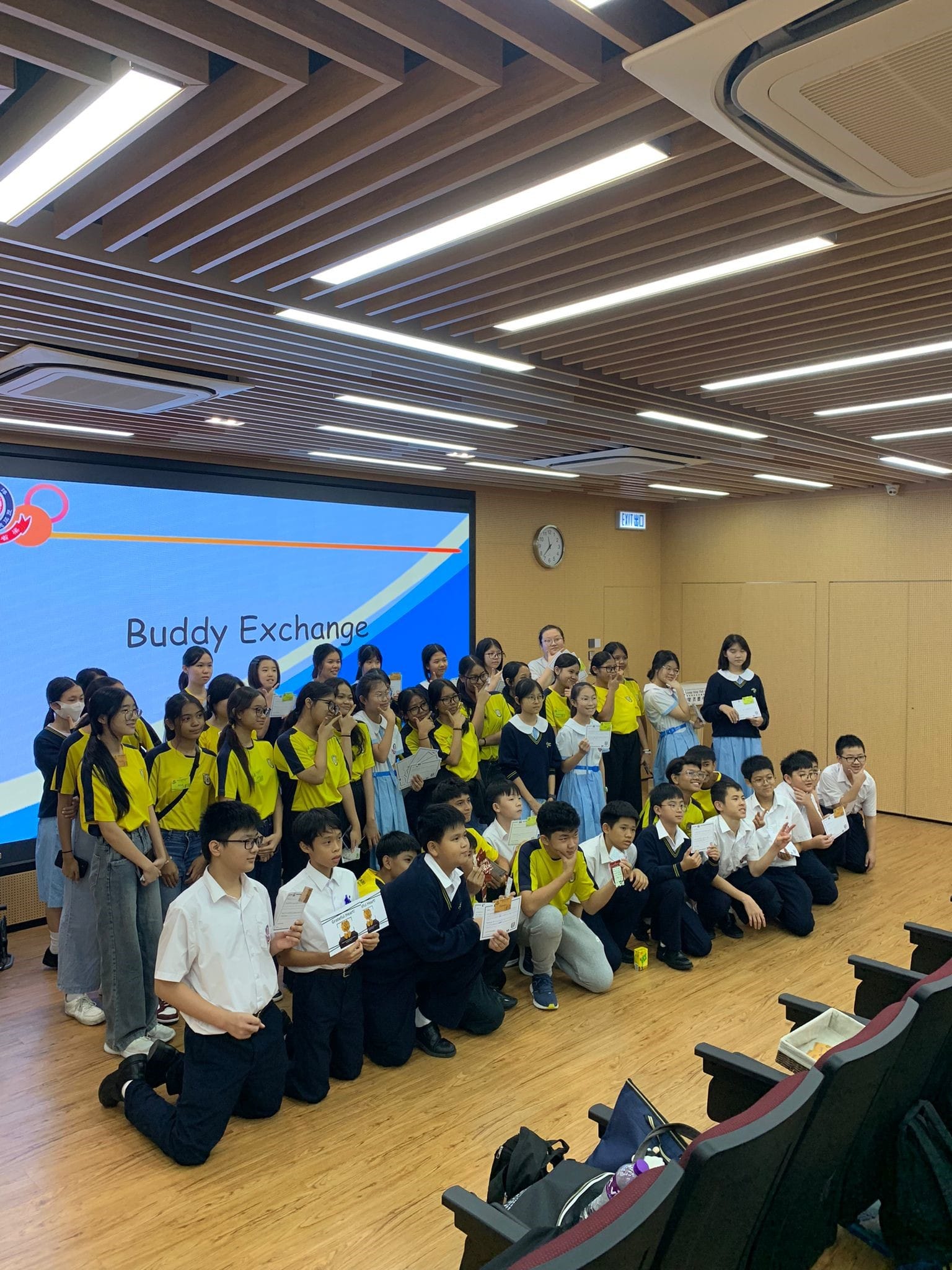 A large group of students in school uniforms pose in a hall in front of a screen displaying "Buddy Exchange."