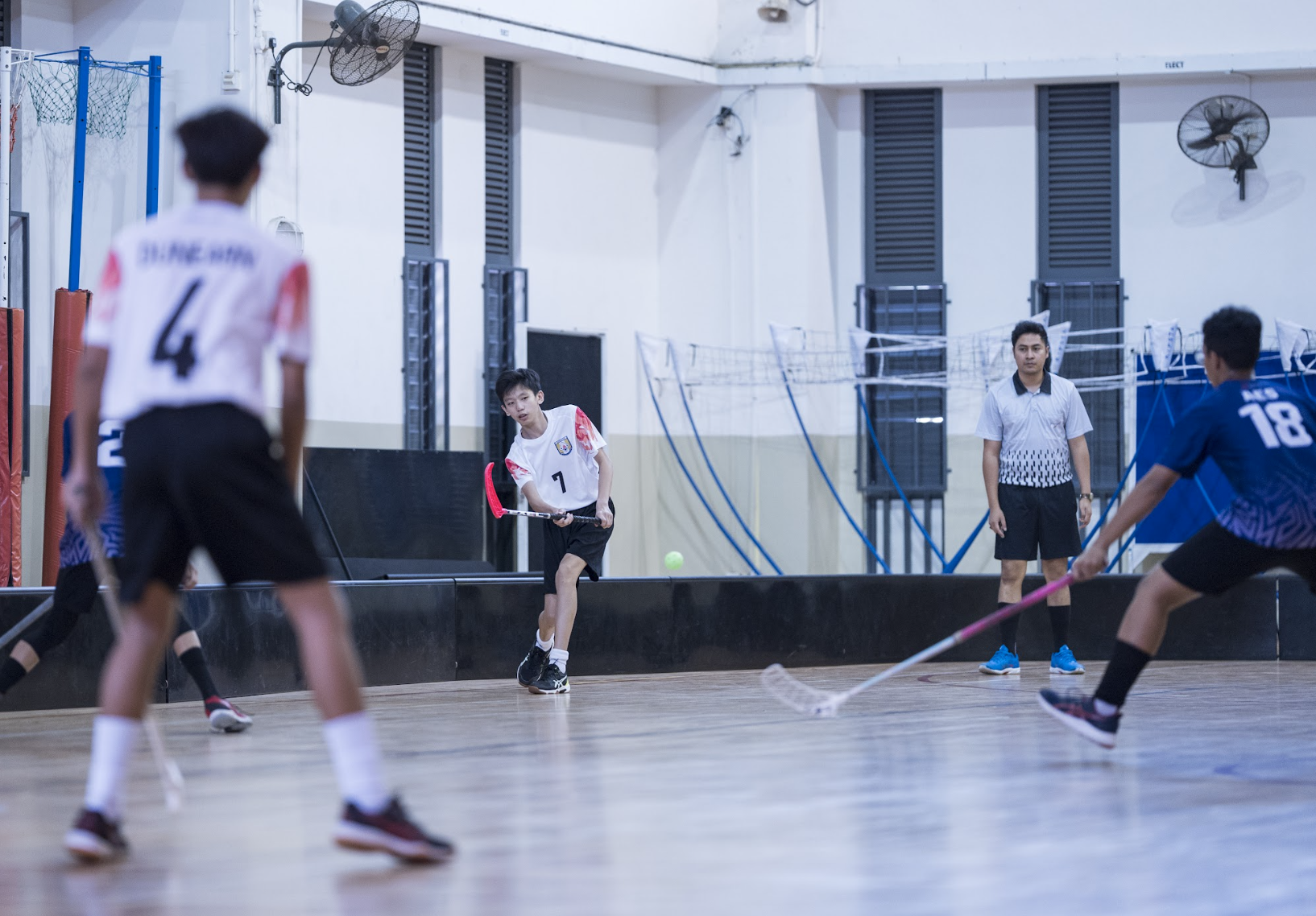 Youth players compete in an indoor floorball game on a wooden court, with an adult referee watching nearby.