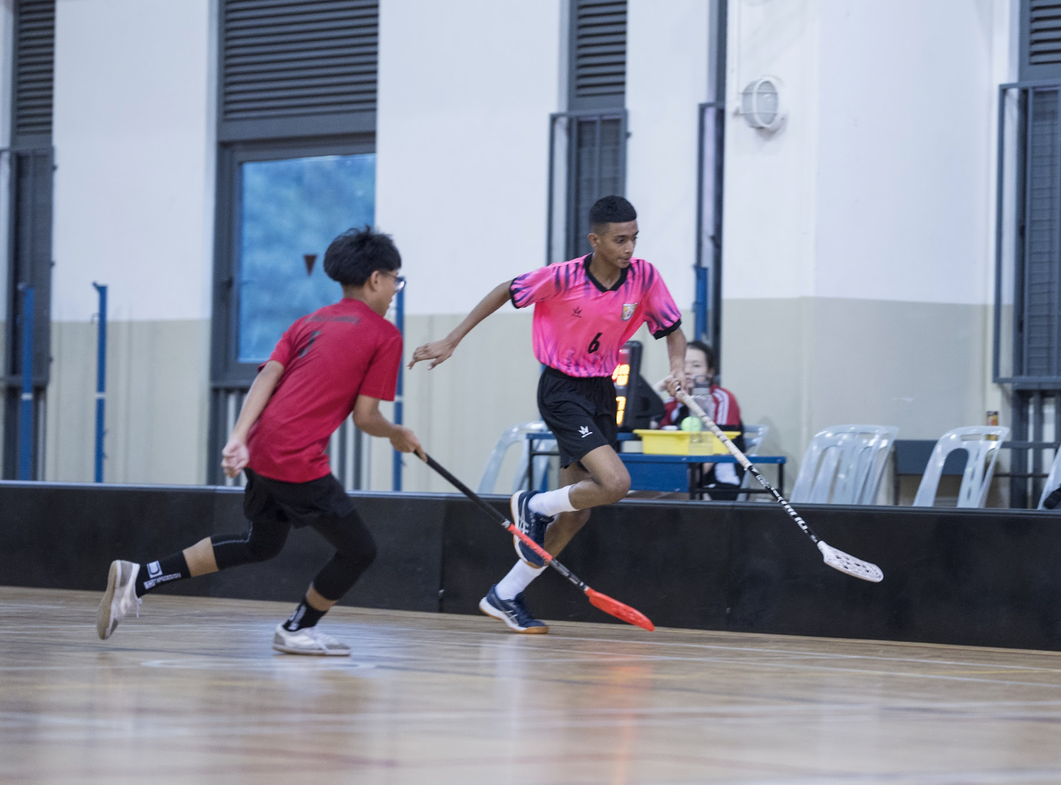 Two young players compete in an indoor floorball match, one in red and one in pink jersey number 6.