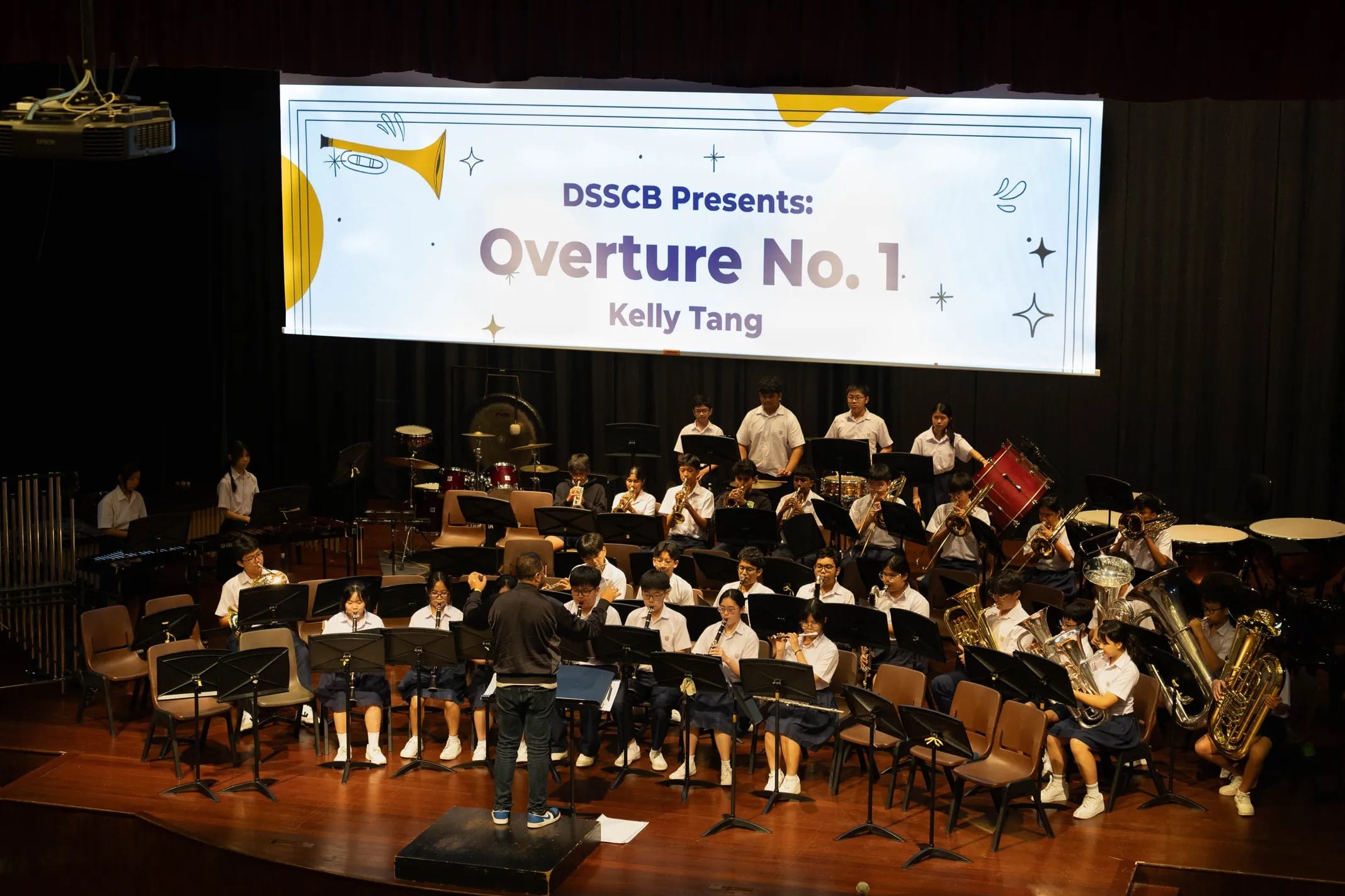 Student band performing on stage with a conductor, and a screen reading DSSCB Presents Overture No. 1 by Kelly Tang.