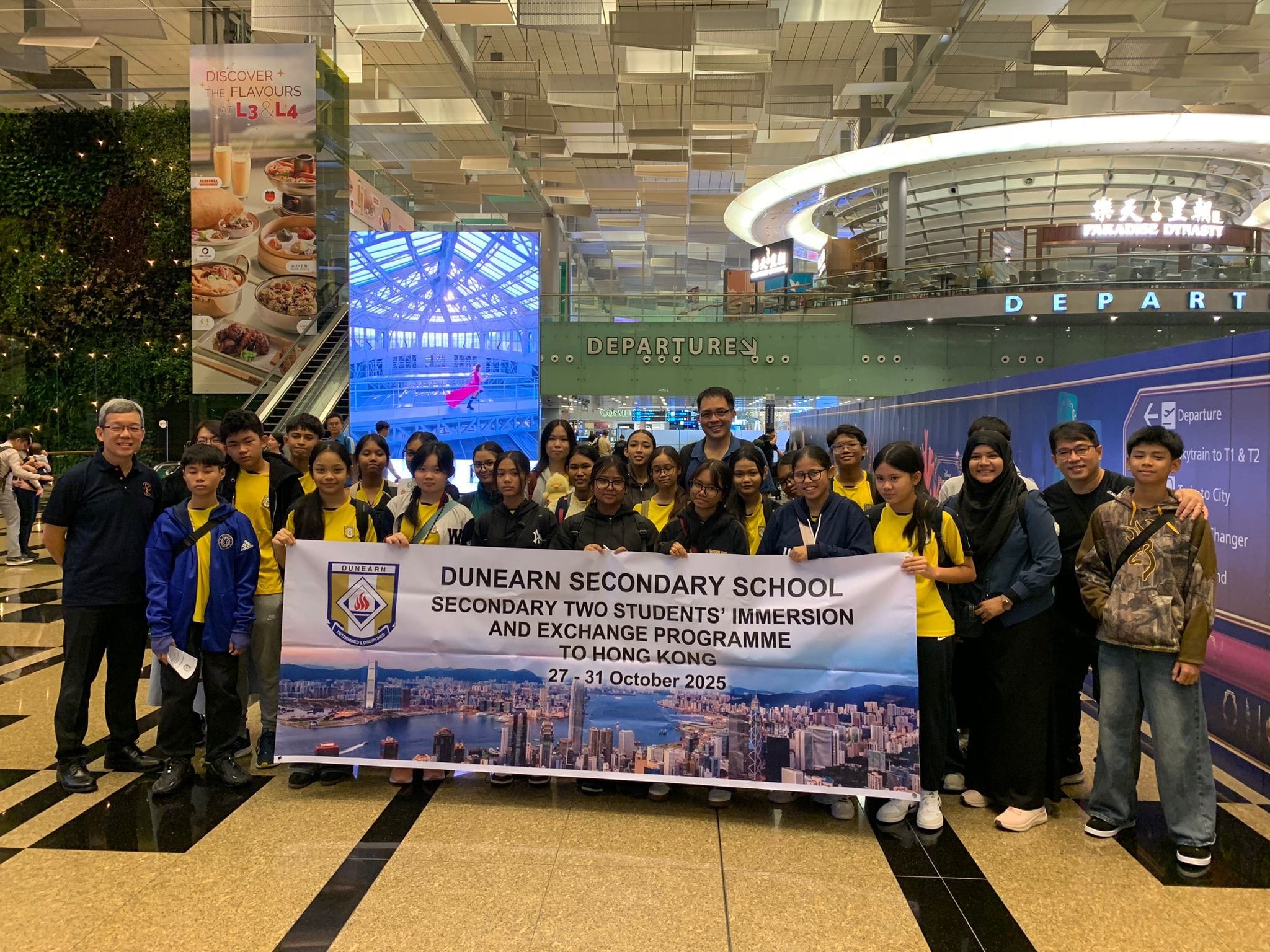 Group of students and teachers at an airport departure hall holding a Dunearn Secondary School Hong Kong exchange programme banner.
