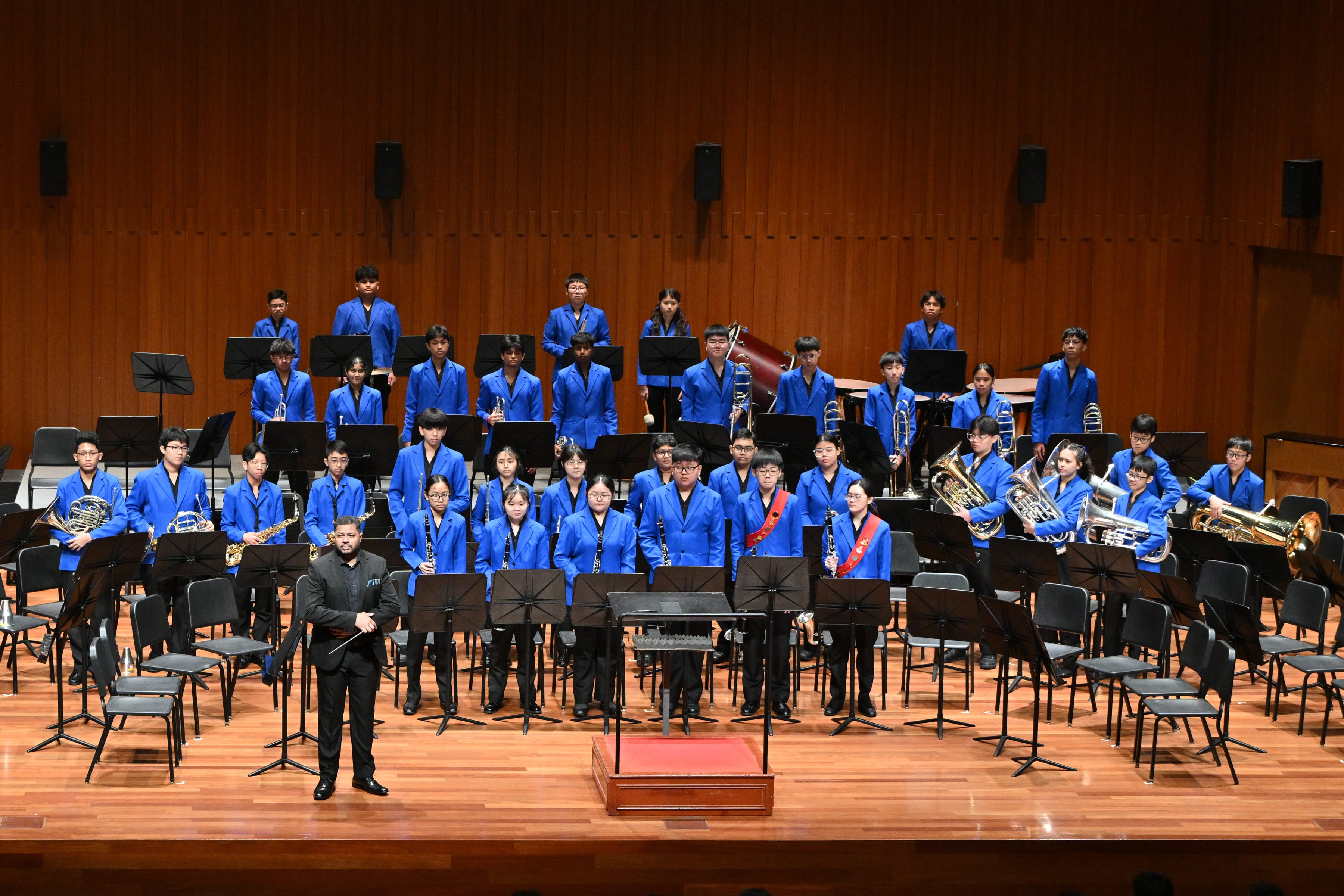 Large student band in blue jackets on a concert stage with instruments, music stands, and a conductor in front.