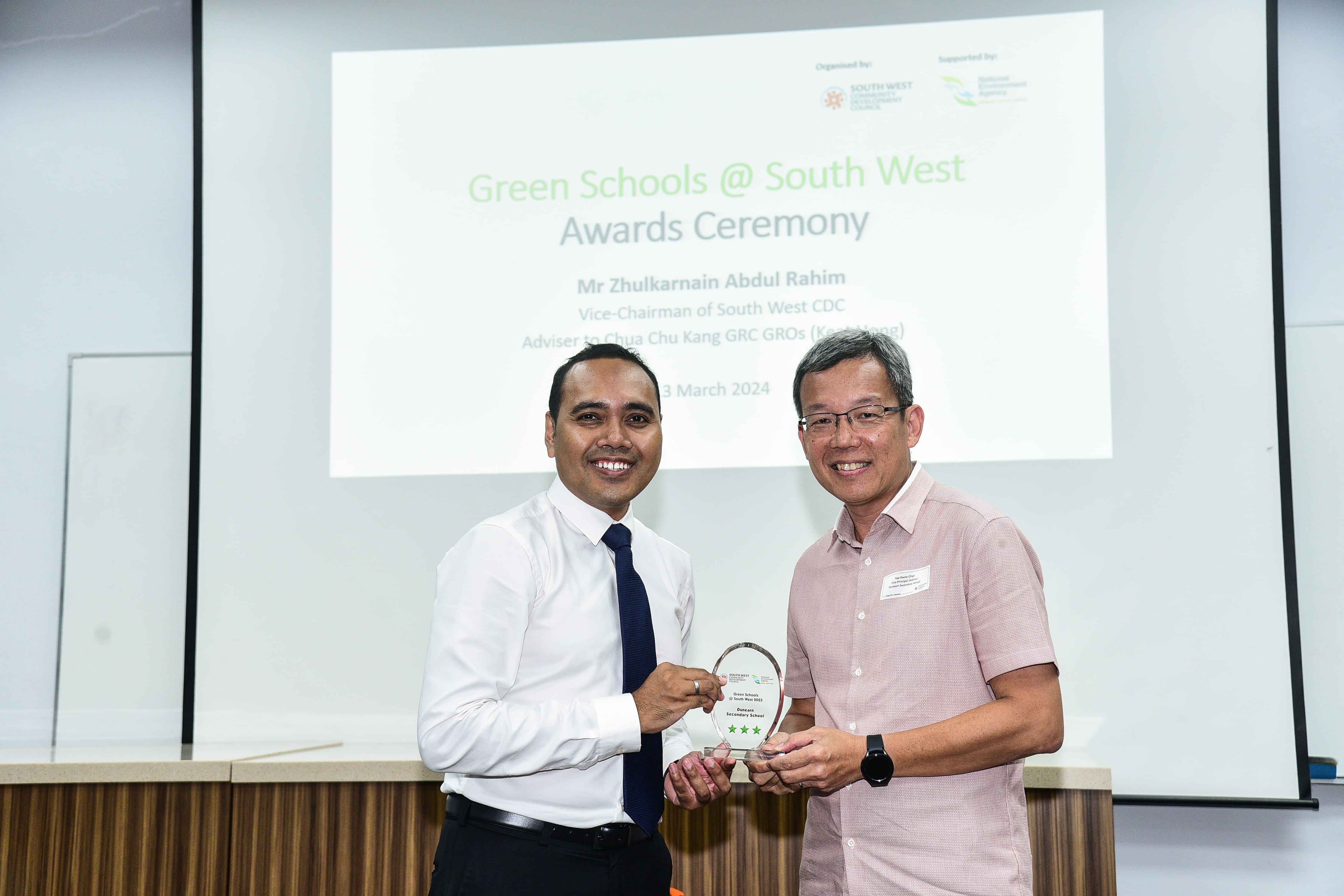 Two men smiling and holding an award plaque, with a projected Green Schools awards ceremony slide behind.