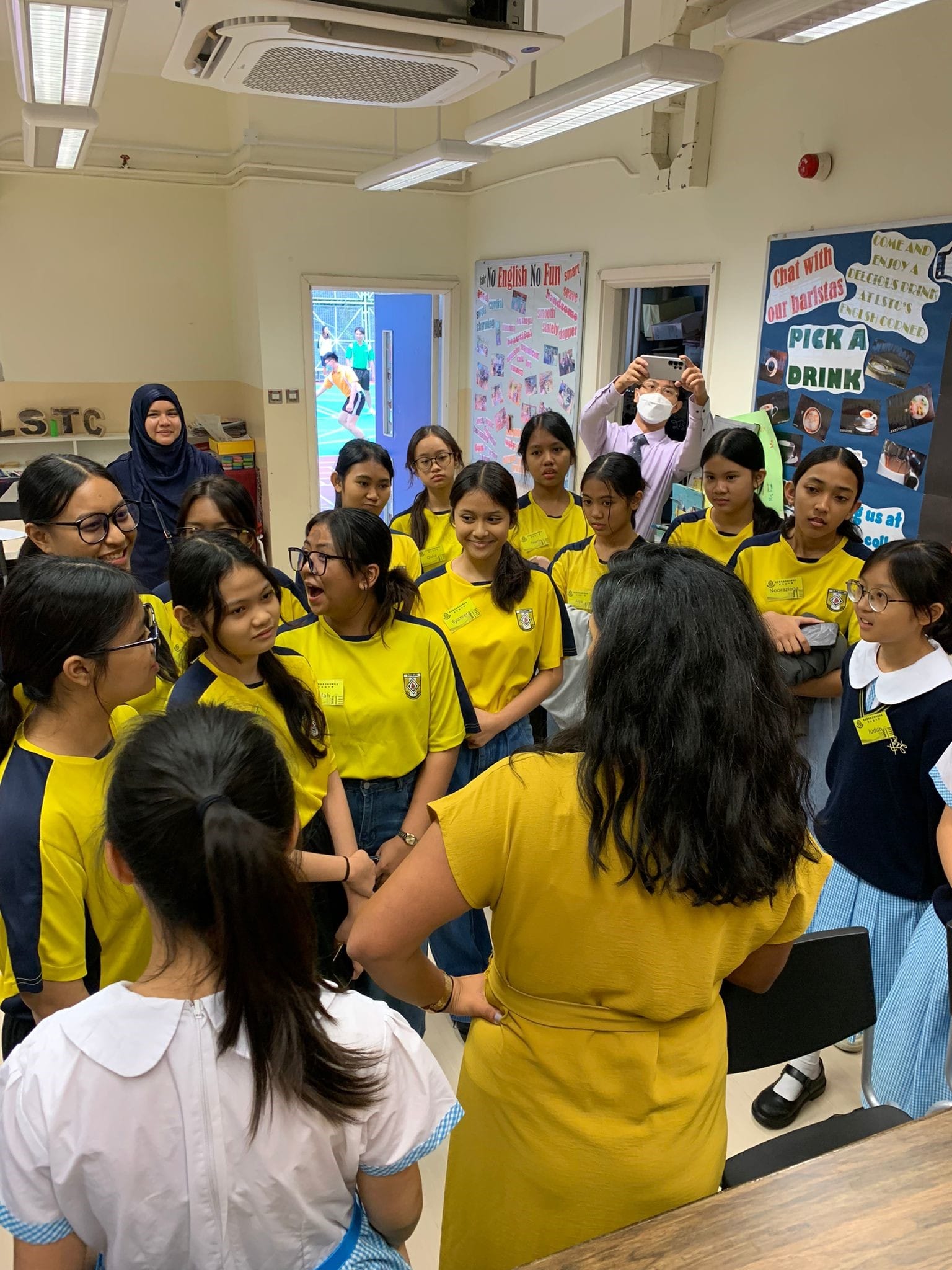 A group of students in yellow uniforms gathers around a teacher in a yellow dress inside a classroom.