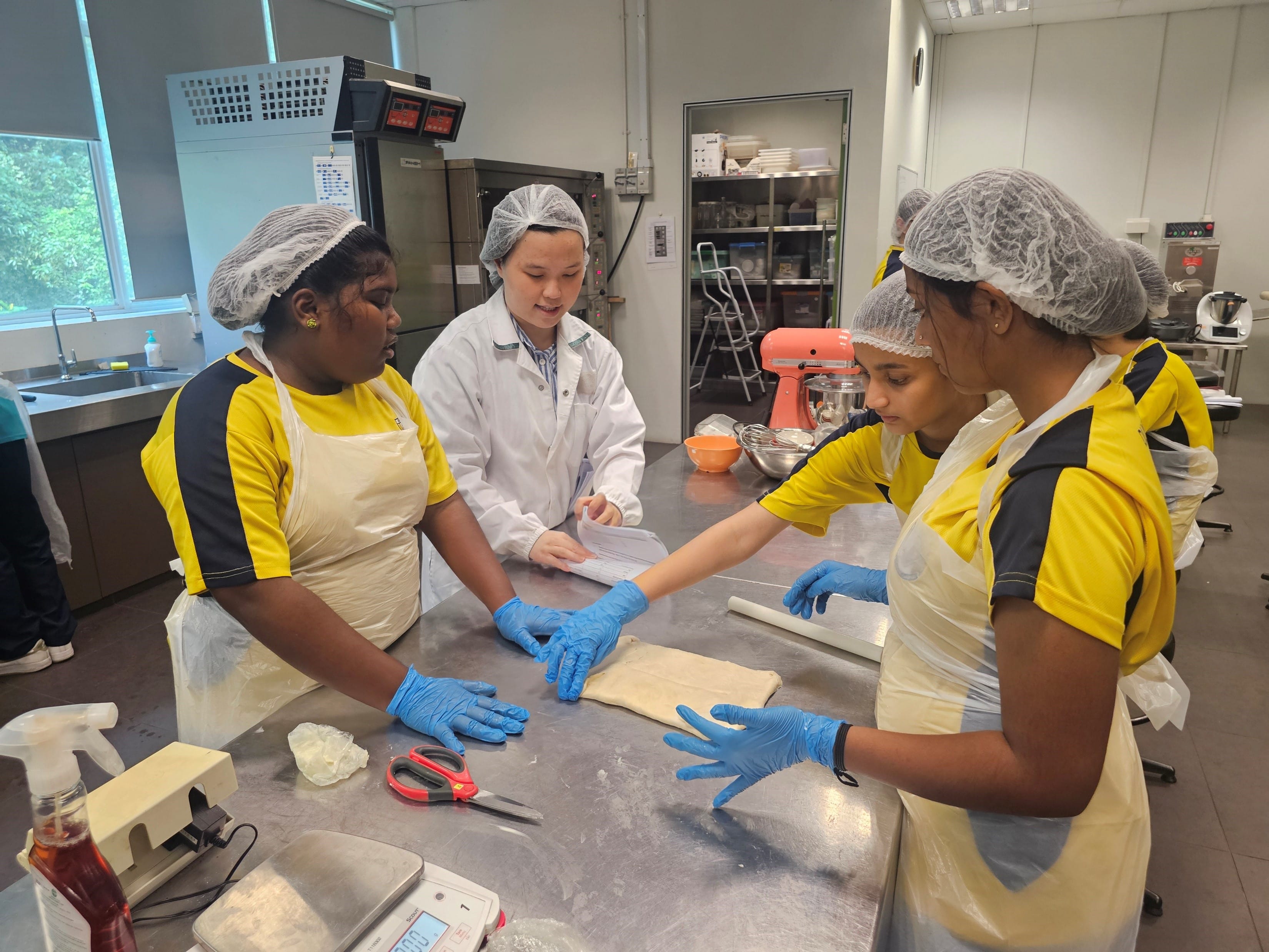 Students in hairnets and blue gloves work with dough at a steel counter in a commercial kitchen, guided by an instructor.