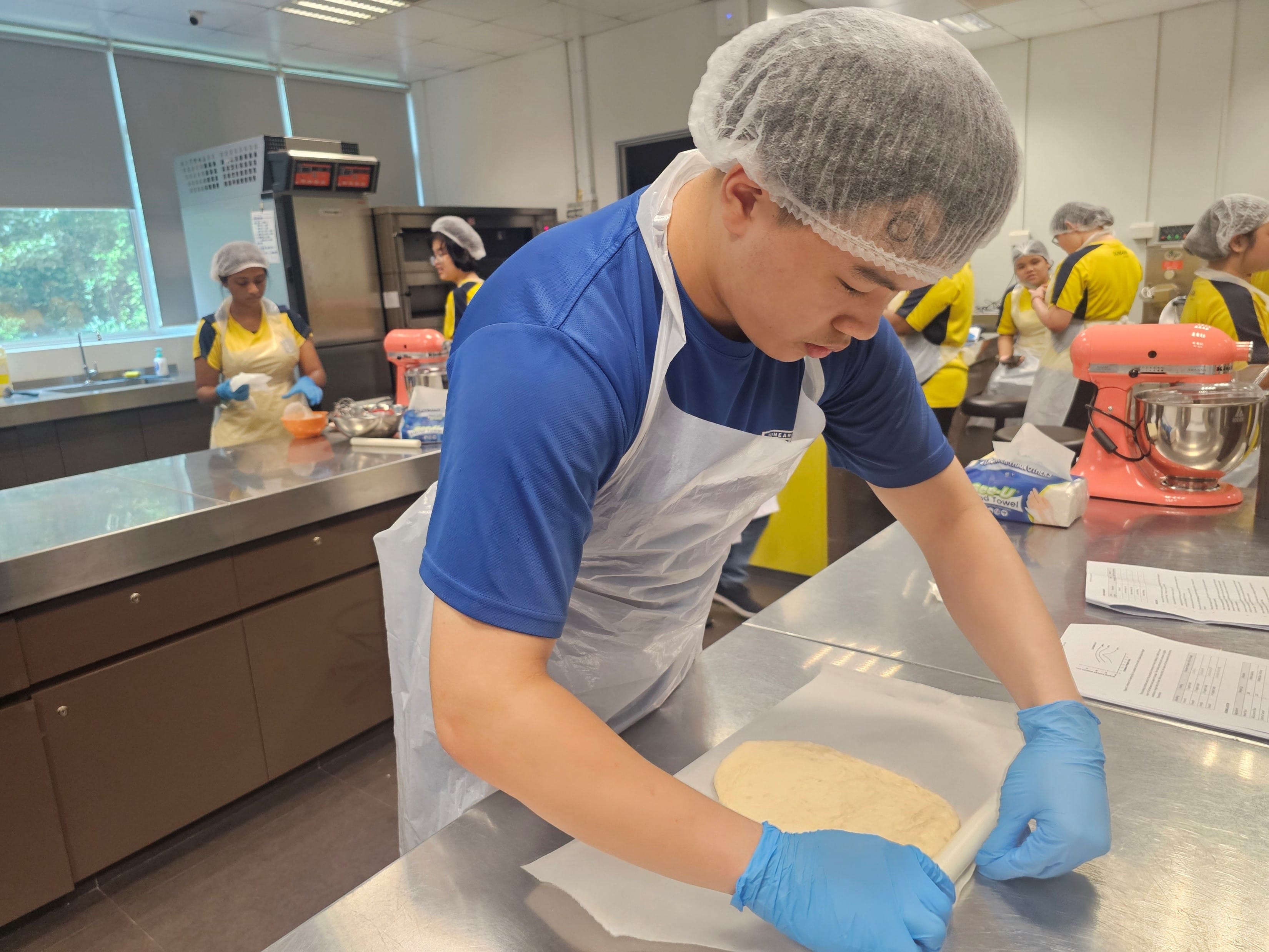 A young person in a hairnet, apron, and blue gloves rolls dough on a steel counter in a kitchen classroom.
