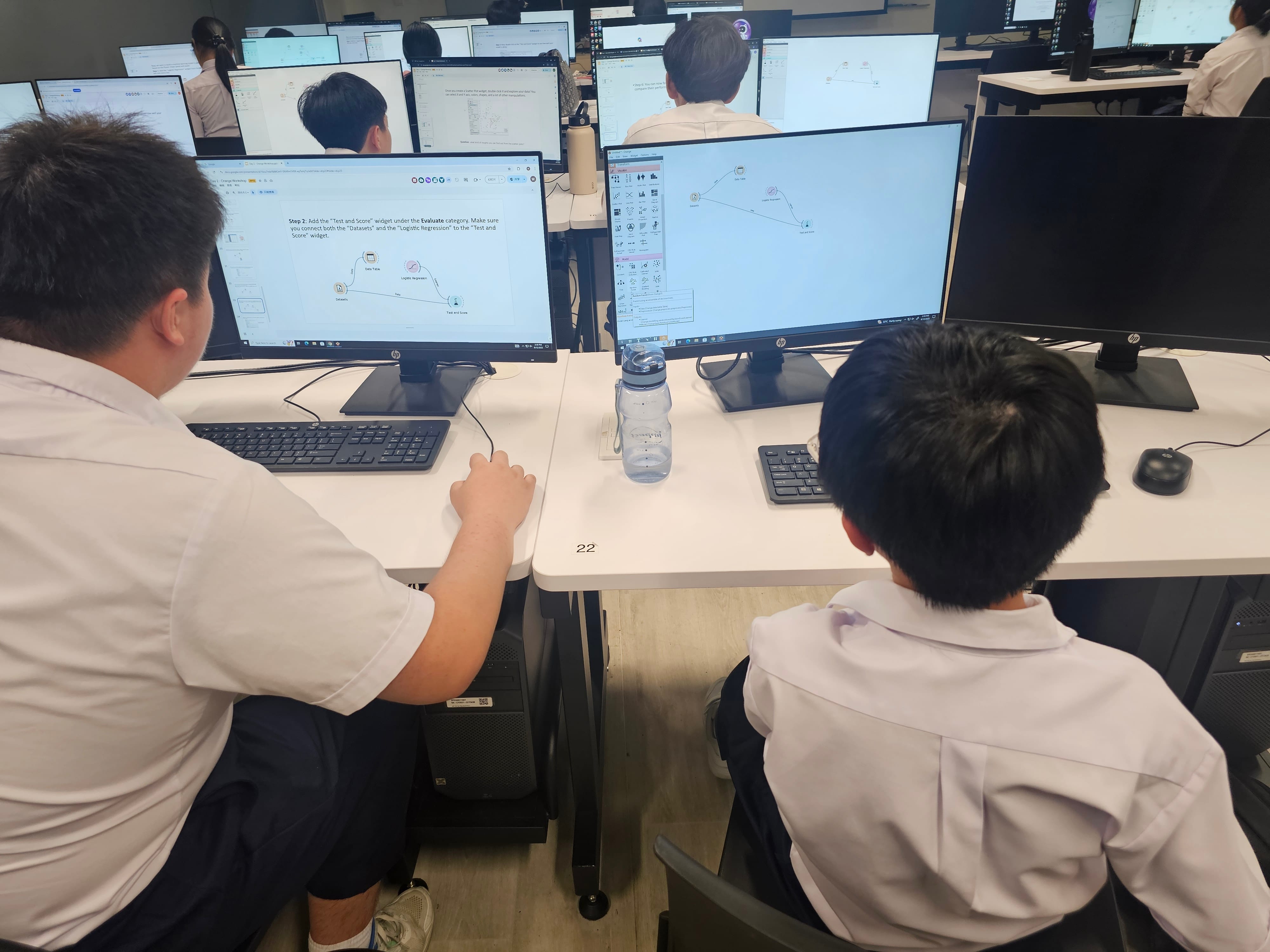 Students in a computer lab work at desktop monitors showing a diagram; a water bottle sits on the desk.