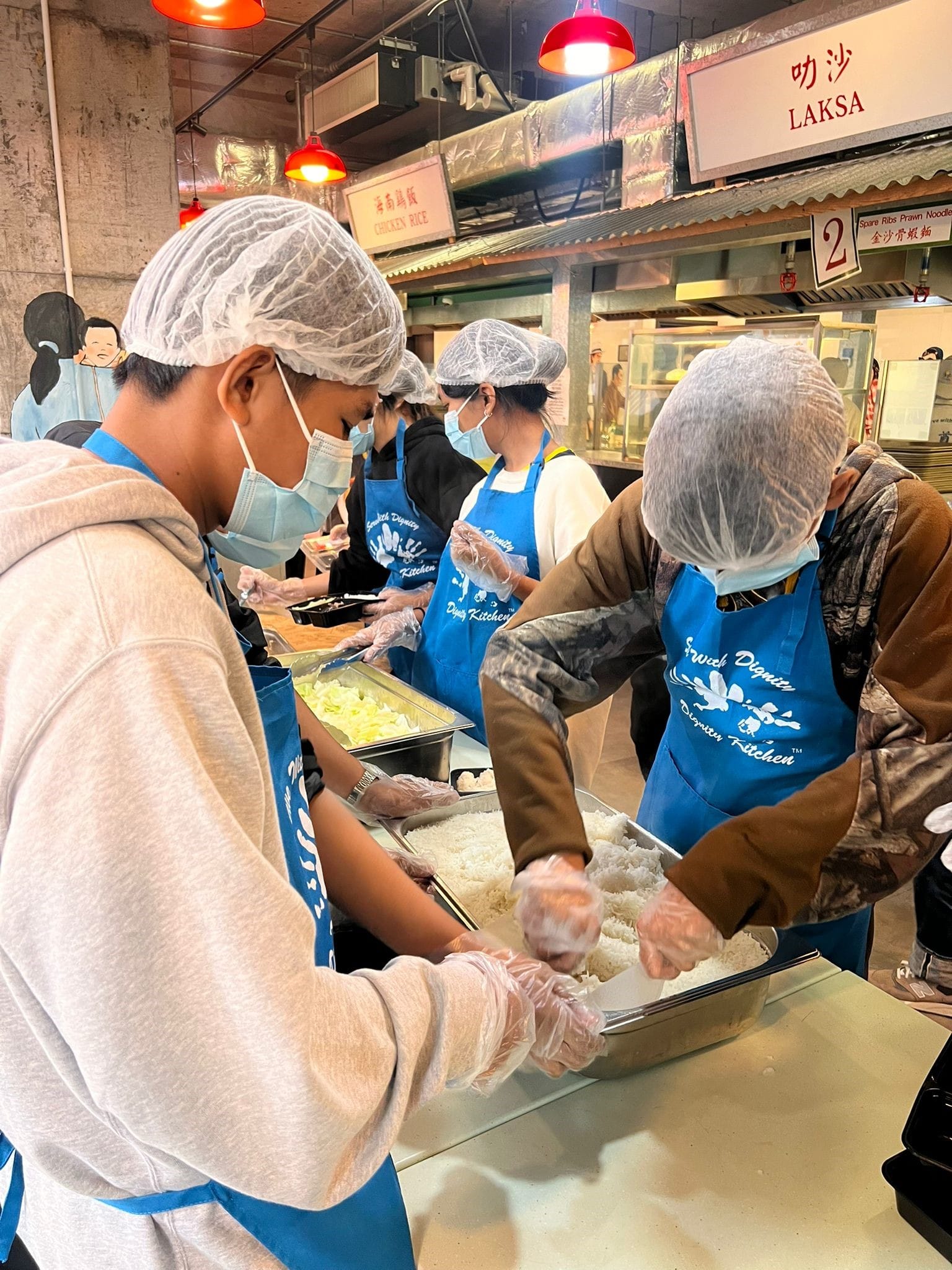 Volunteers in blue aprons, hairnets, and masks serving rice at a food kitchen with Laksa signs overhead.