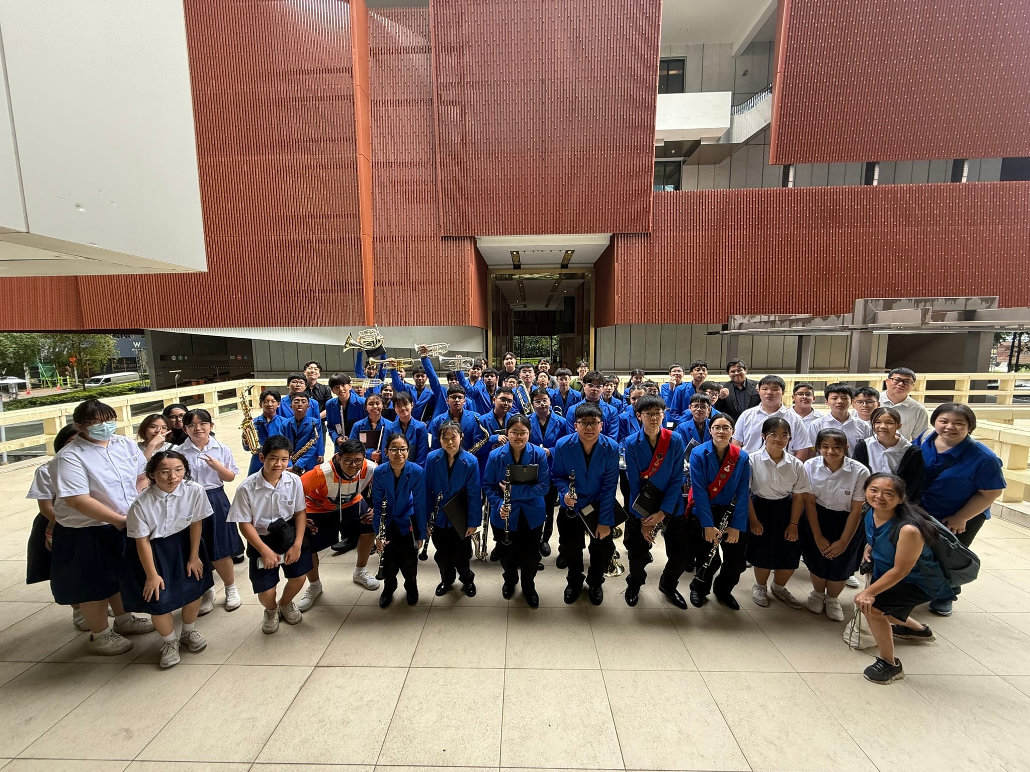 A large group of students in blue uniforms holding band instruments pose outdoors in front of a red building.