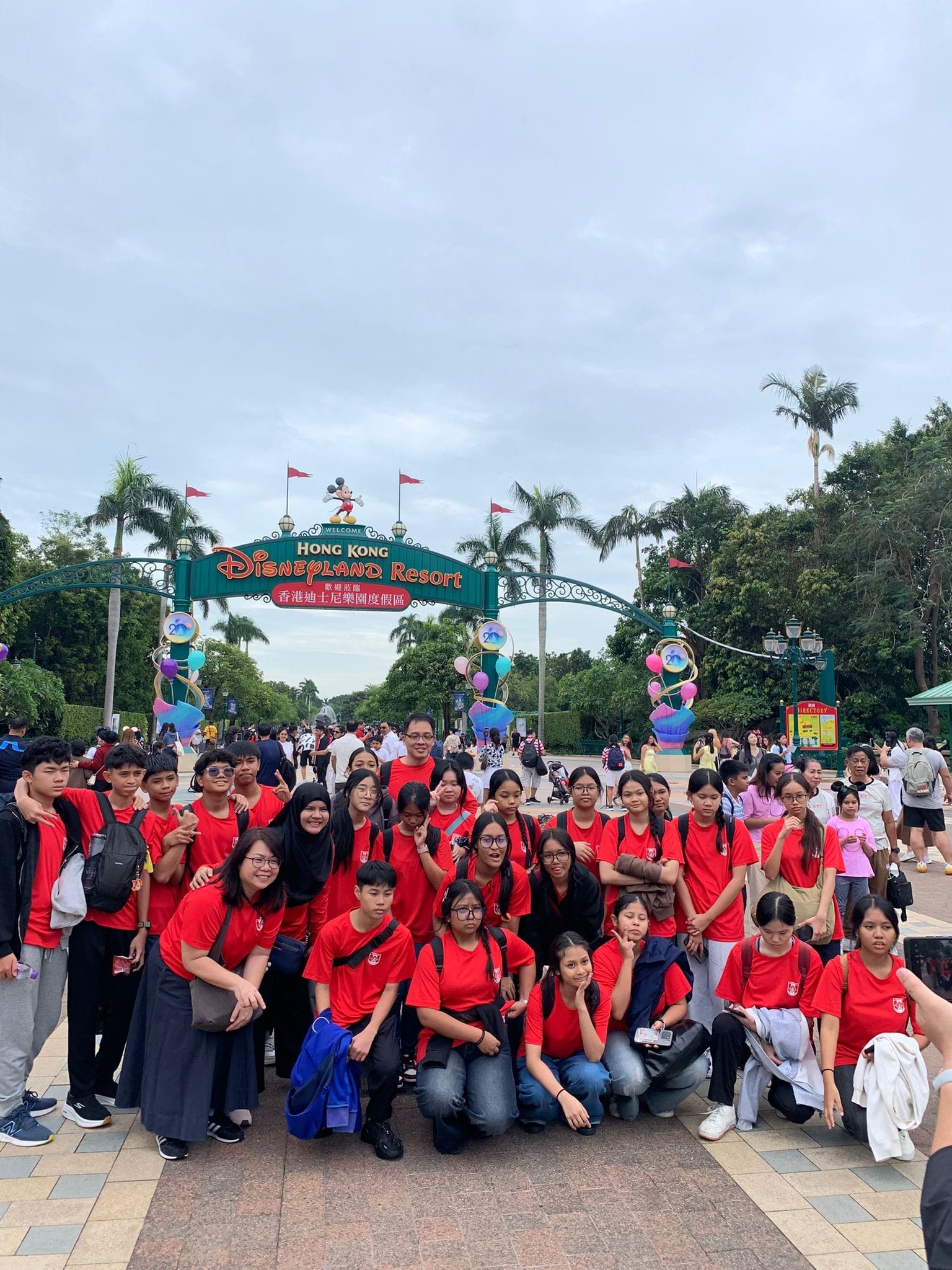 A large group of students in red shirts poses in front of the Hong Kong Disneyland Resort entrance sign.