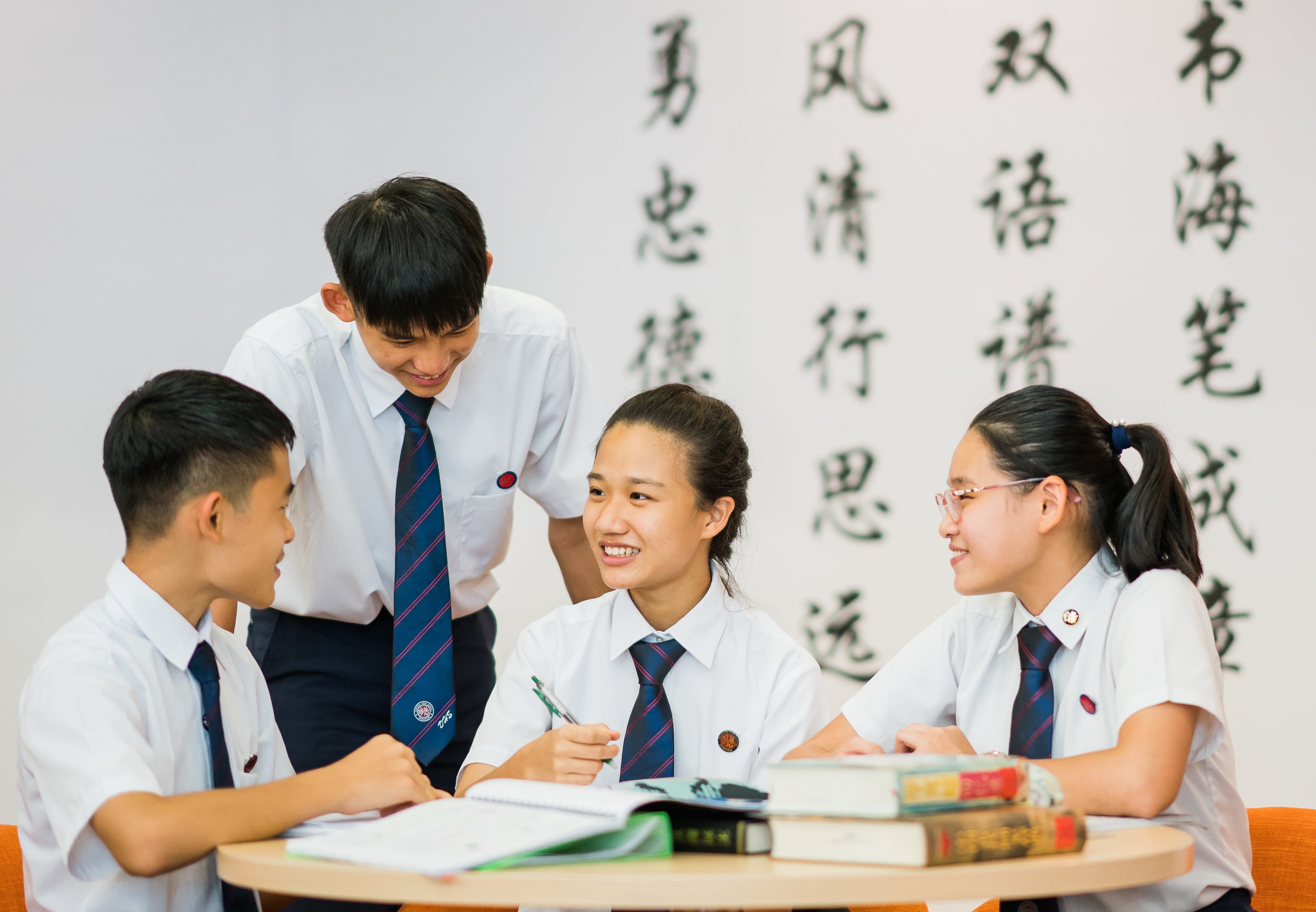 Four students in white uniforms and striped ties study together at a table with books, smiling, near a wall with Chinese calligraphy.