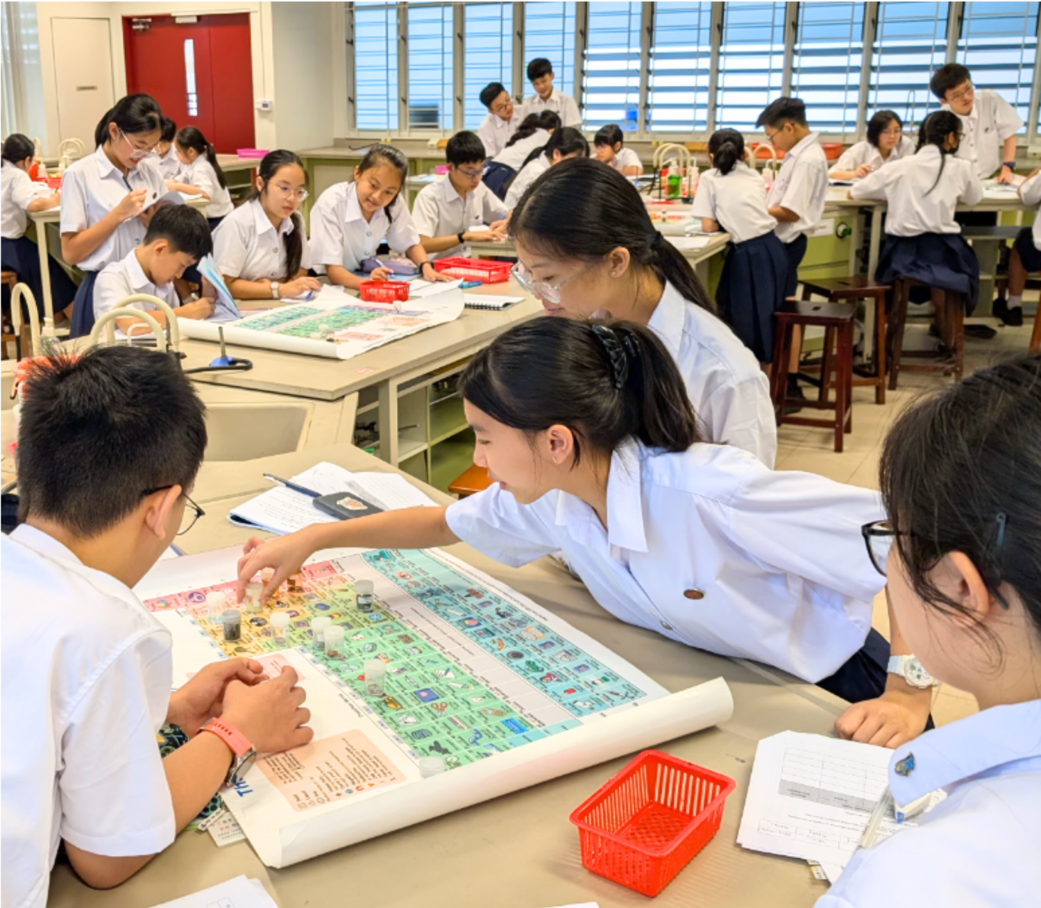 Students in white uniforms work together around a large periodic table poster on a desk in a science classroom.