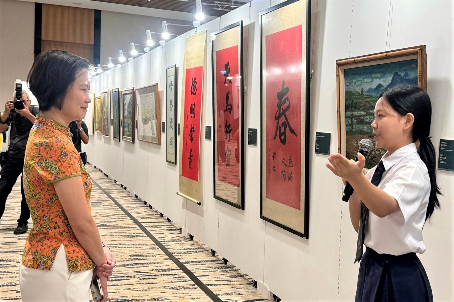 A schoolgirl holding a microphone speaks to a woman in an orange top at a Chinese calligraphy art exhibition.