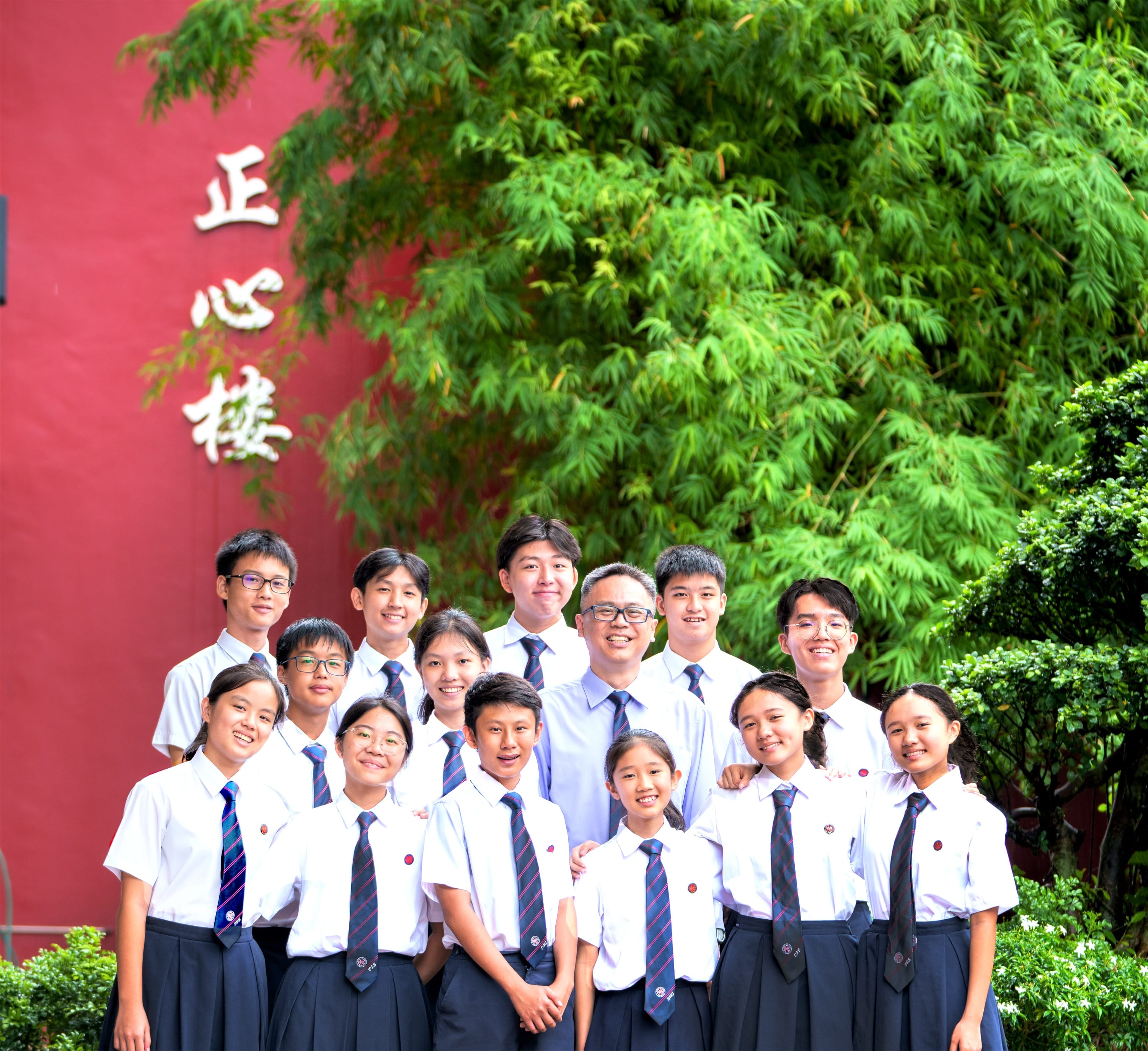 A group of students in white shirts and striped ties pose smiling before a red wall with Chinese characters and green trees.