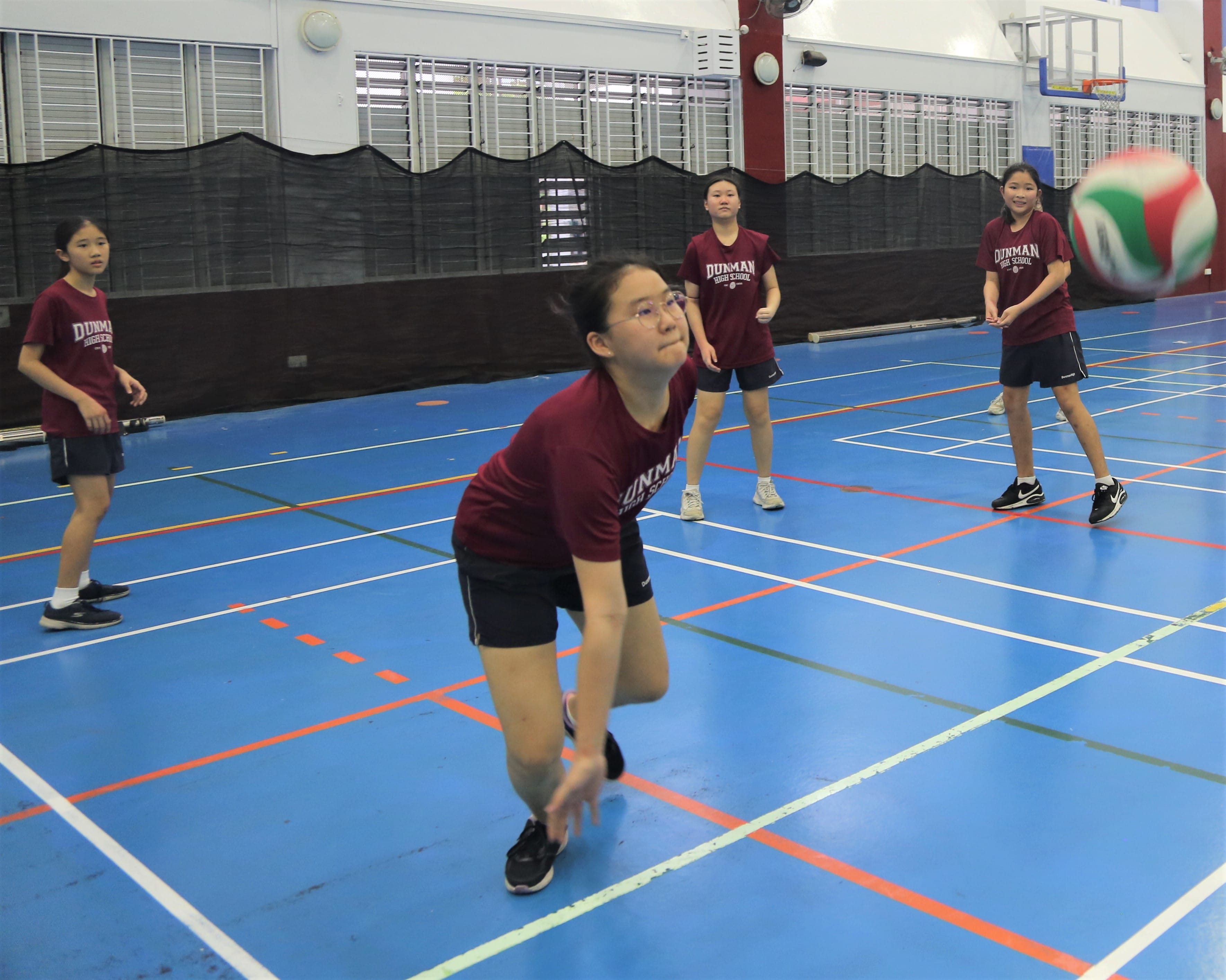 Four girls in maroon Dunman High School shirts practice volleyball on a blue indoor court, one diving to receive the ball.