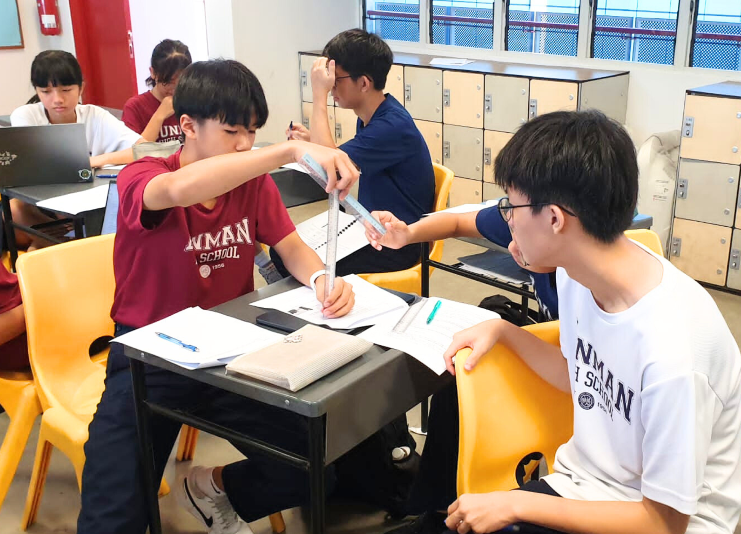 Two students share a ruler at a desk while classmates work nearby in a classroom with yellow chairs and lockers.