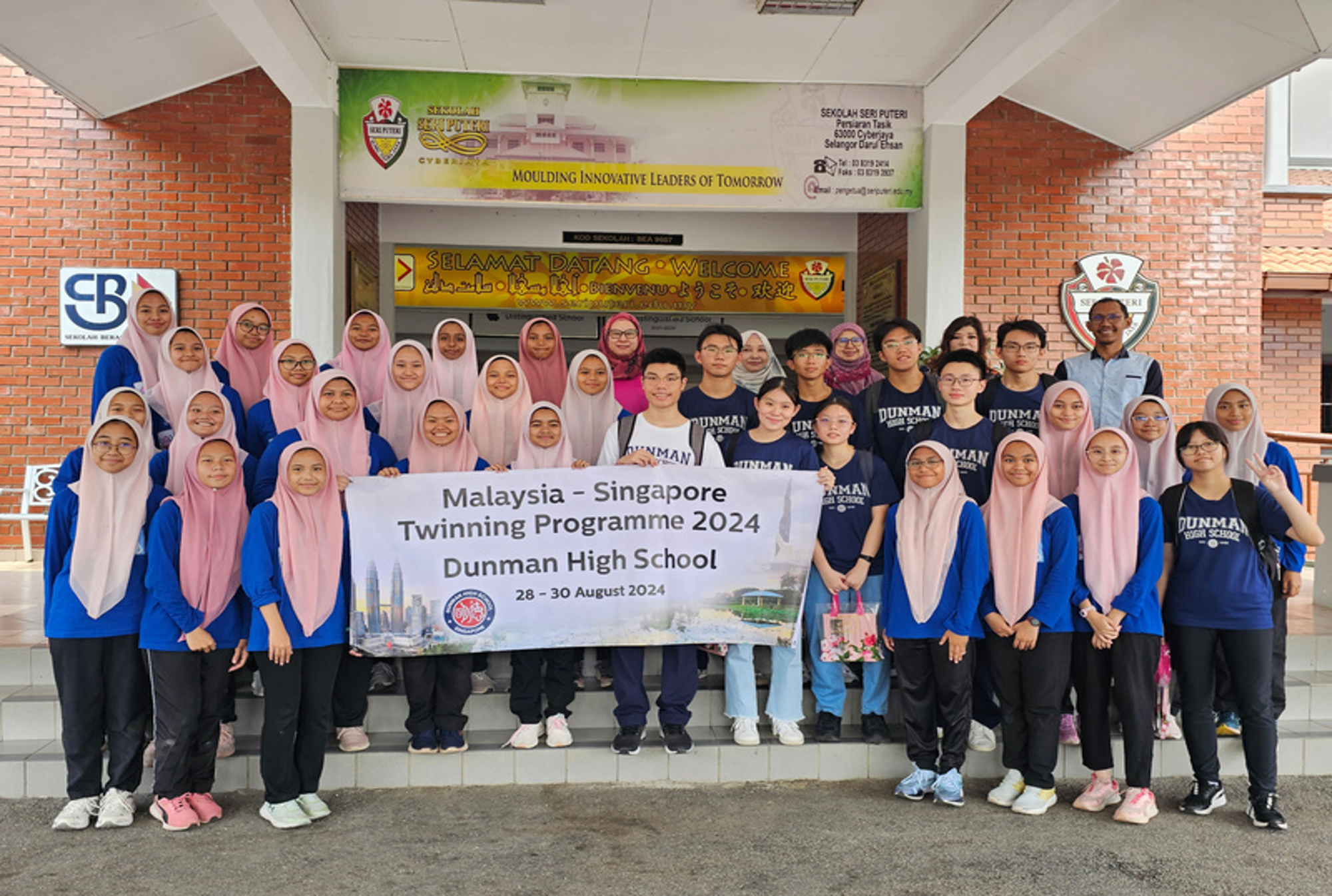 Group of students in front of a school entrance holding a Malaysia–Singapore Twinning Programme 2024 banner.