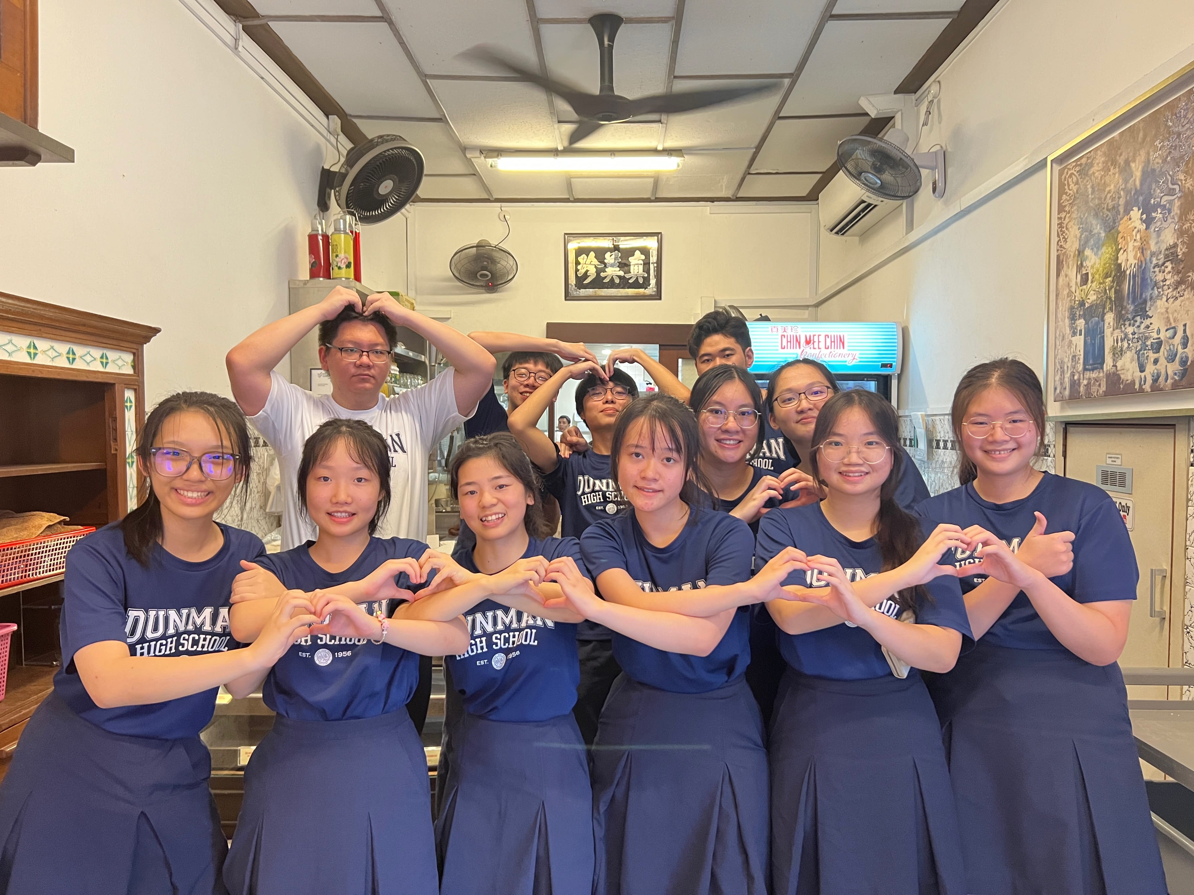 A group of Dunman High School students in navy uniforms pose making heart shapes with their hands indoors.