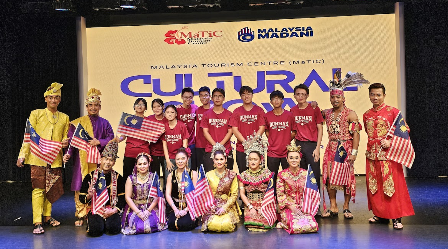 Group of people in traditional Malaysian costumes and school shirts pose with Malaysian flags at MaTiC Cultural Show.