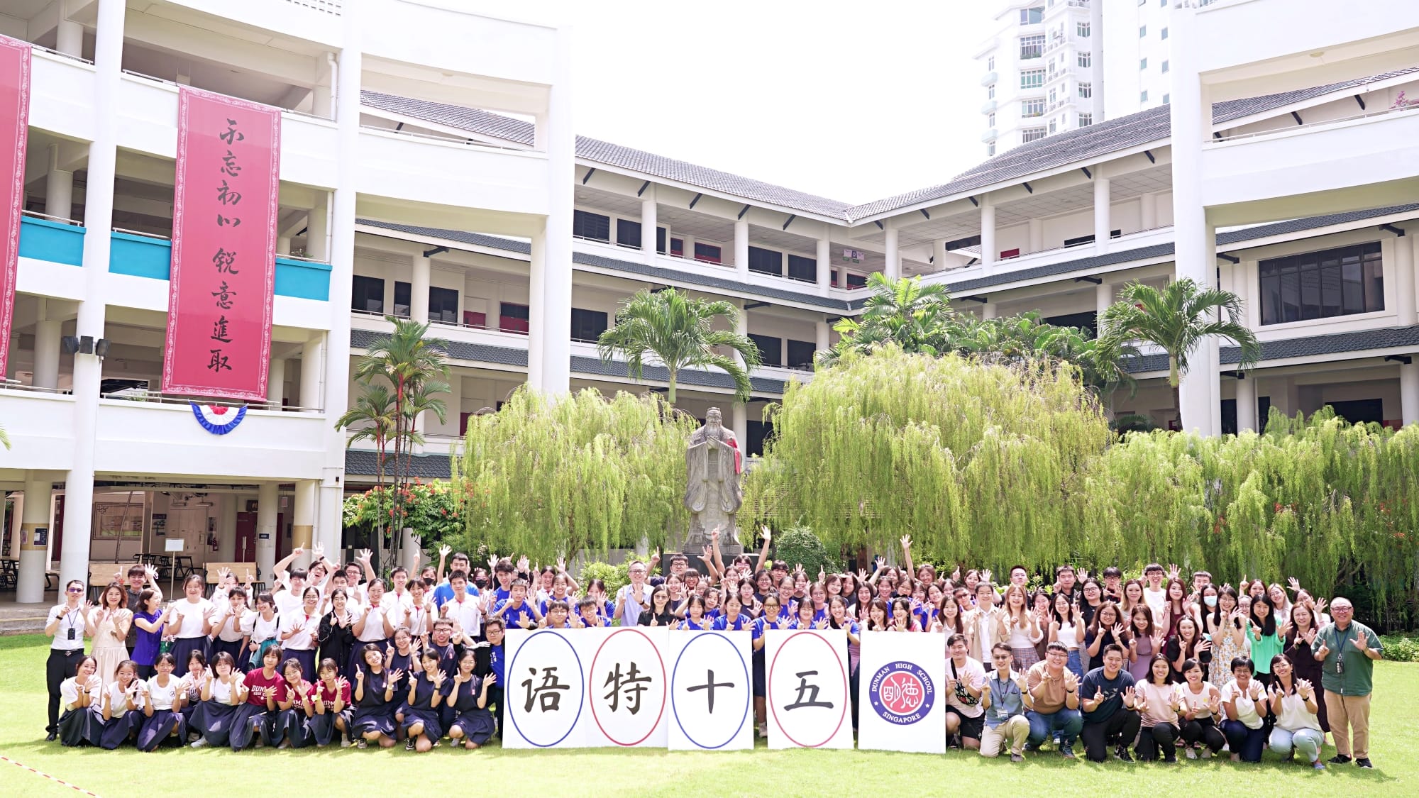 Large group of students posing outdoors at a school courtyard, holding signs with Chinese characters, waving and smiling.