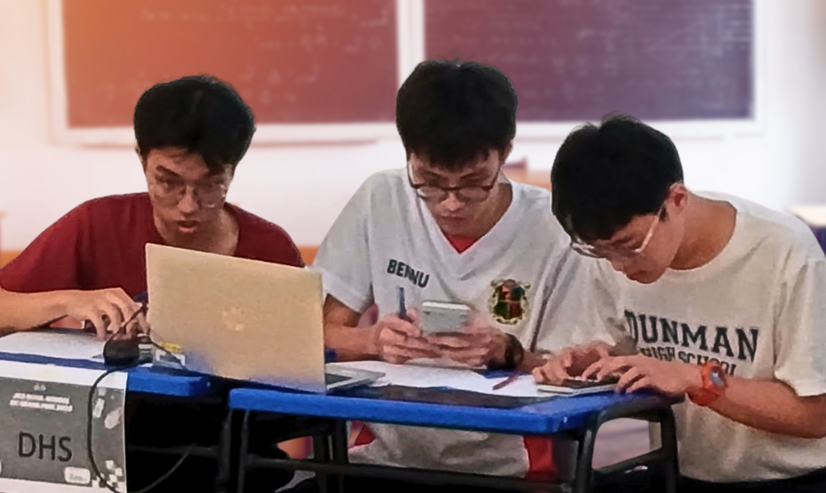 Three teenage boys wearing glasses sit at a desk using a laptop, phone, and tablet in a classroom.