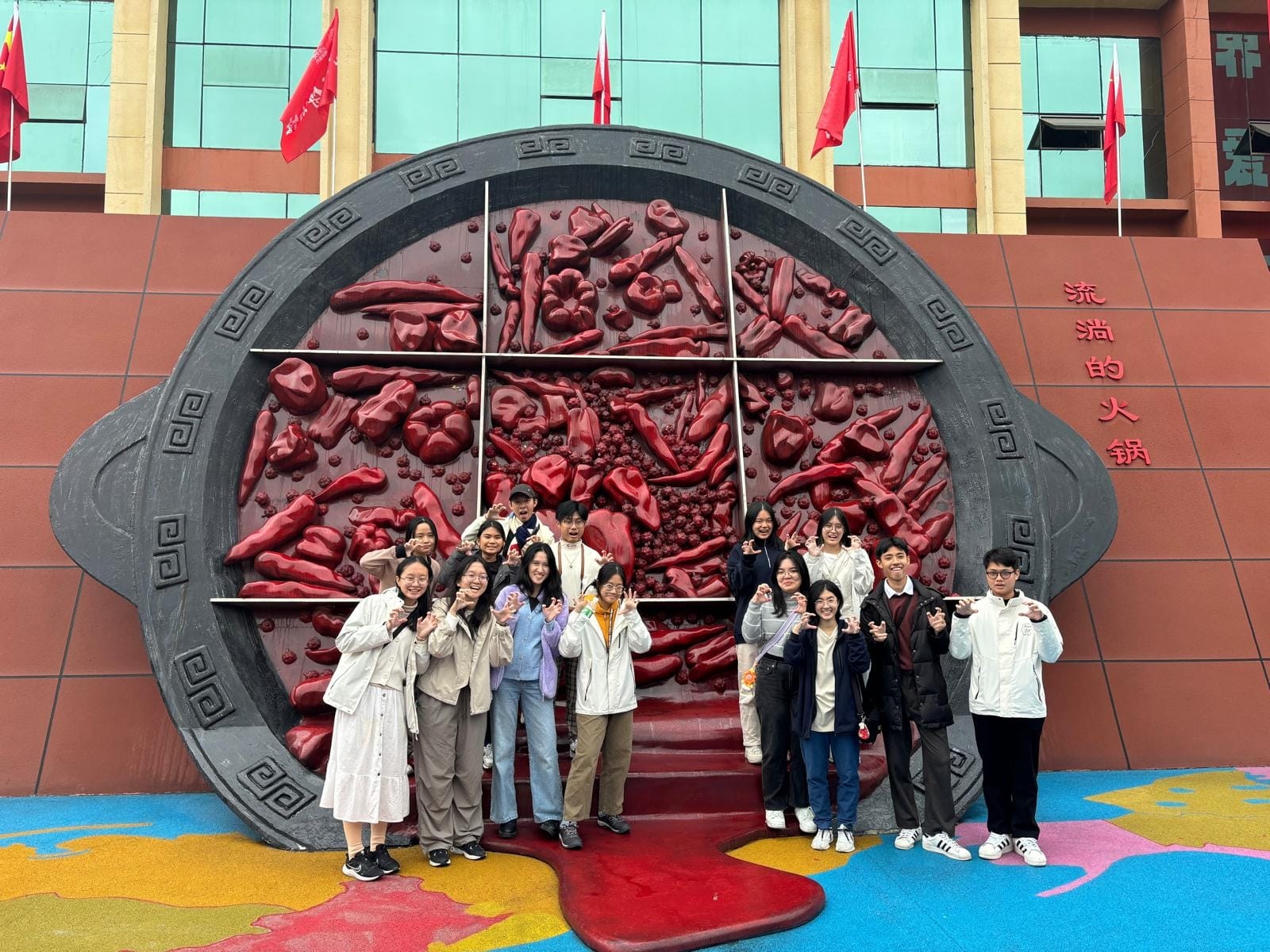 A group of young people posing in front of a large red hotpot sculpture with Chinese characters on a building.