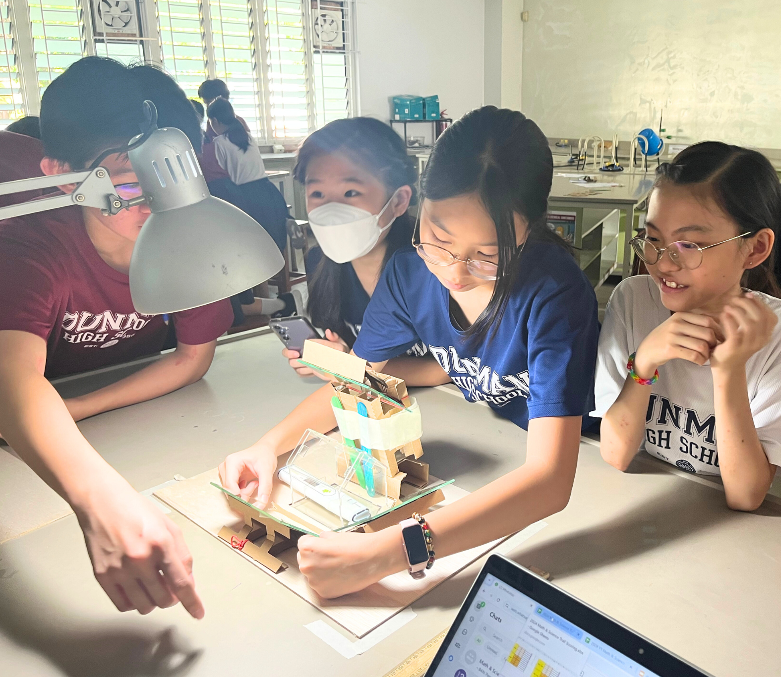 Three students examine a small cardboard and glass model structure on a desk in a classroom.