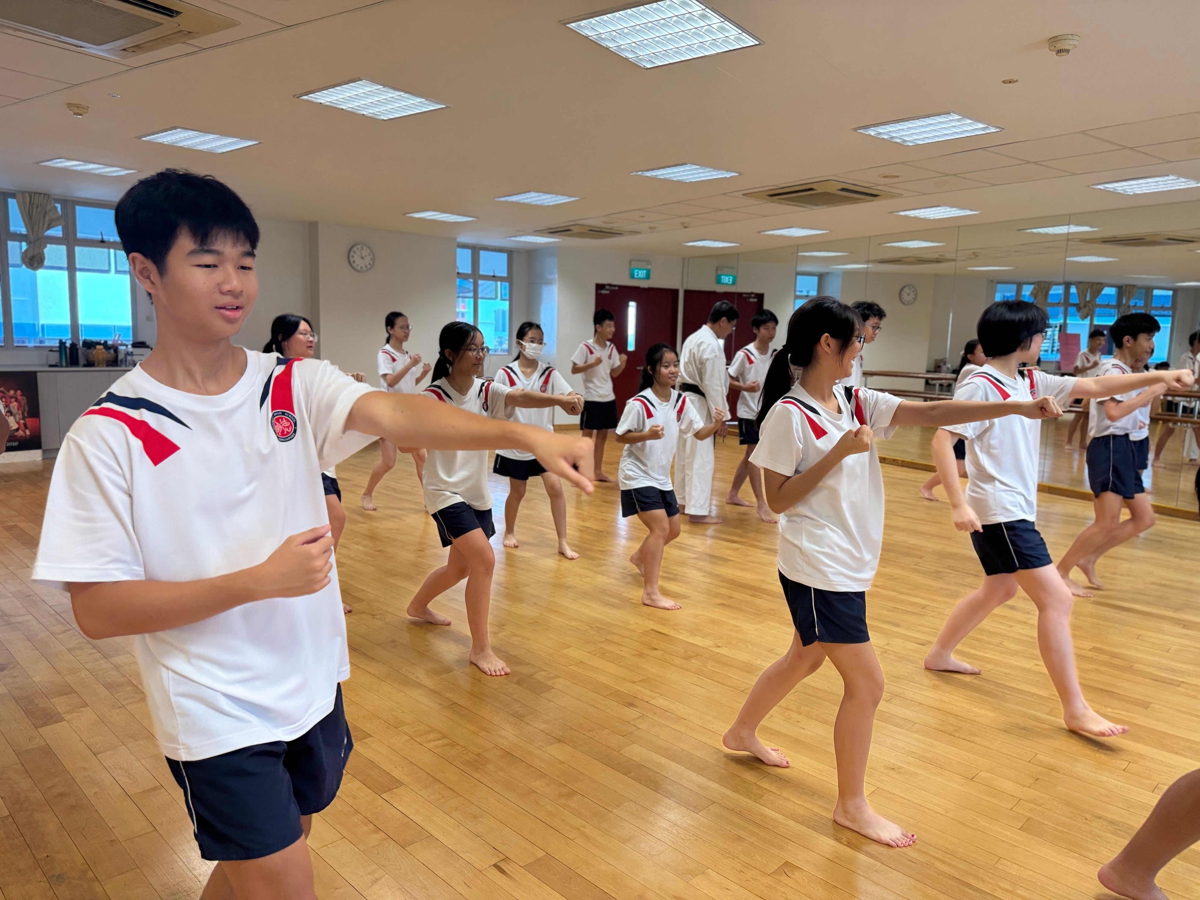 Students in white and navy uniforms practice martial arts moves barefoot in a mirrored studio with wooden floors.